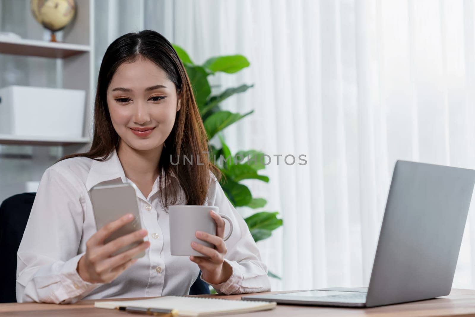 Modern and young enthusiastic businesswoman multitasks by checking her smartphone for business matters and working with laptop in her desk at the office.