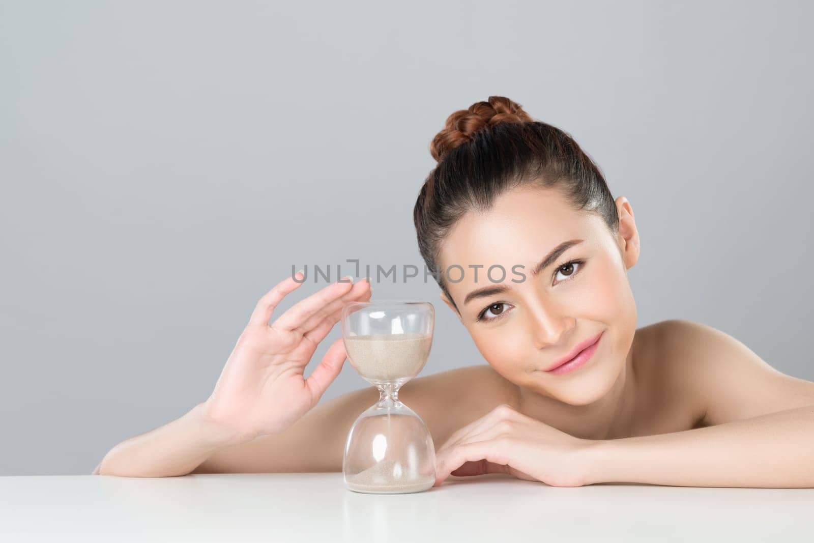 Glamorous model holding hourglass in beauty concept of anti-aging skincare treatment for woman. Young asian girl portrait with perfect smooth clean skin and flawless soft makeup in isolated background