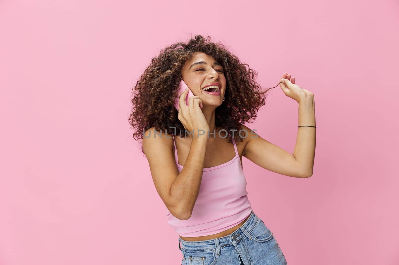 Woman with curly afro hair talking in pink top and jeans on pink background, smile, happiness, copy space. High quality photo