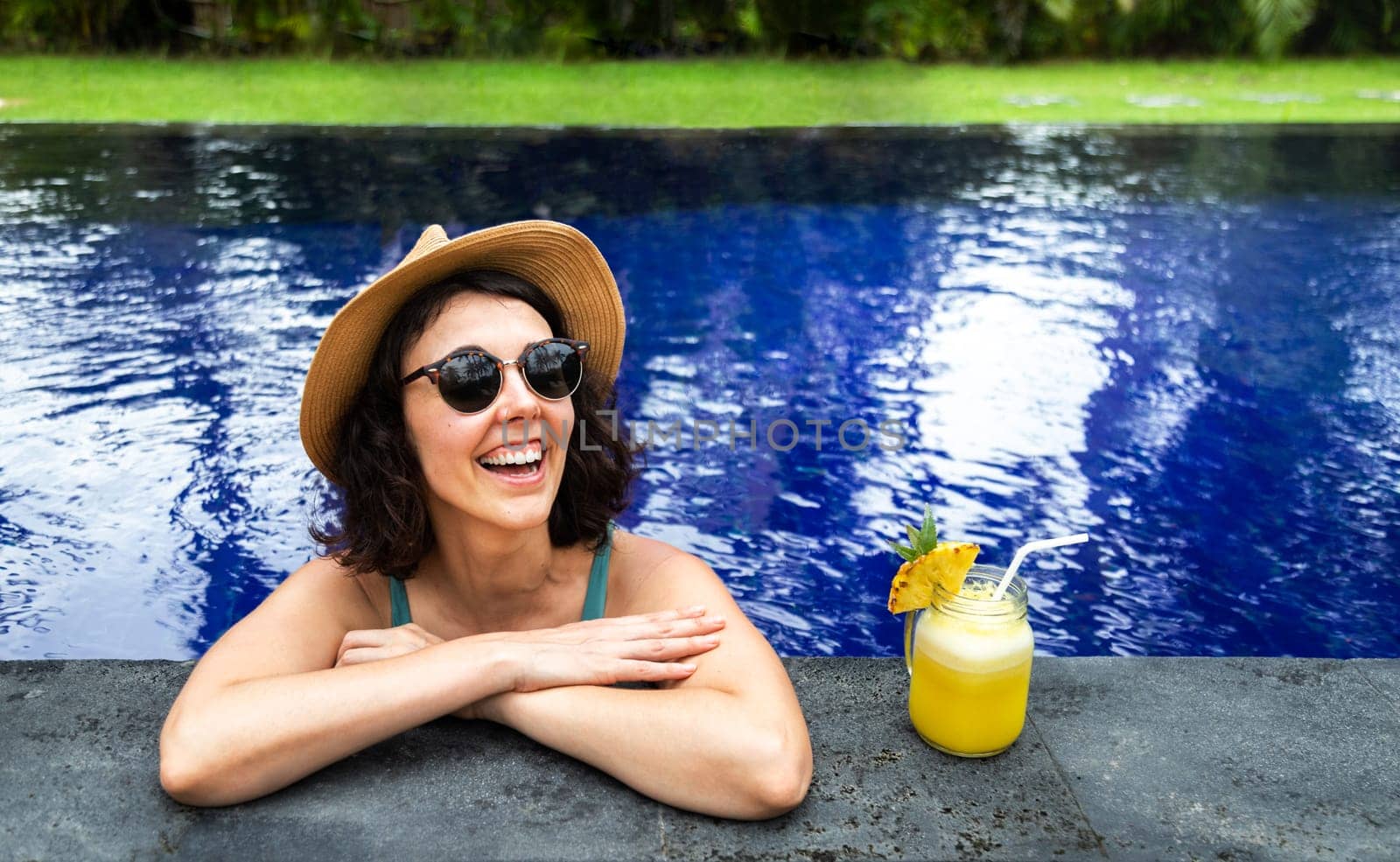Happy young woman relaxing in the swimming pool during summer vacation in tropical resort. Female on holidays drinking healthy juice. Holiday concept.
