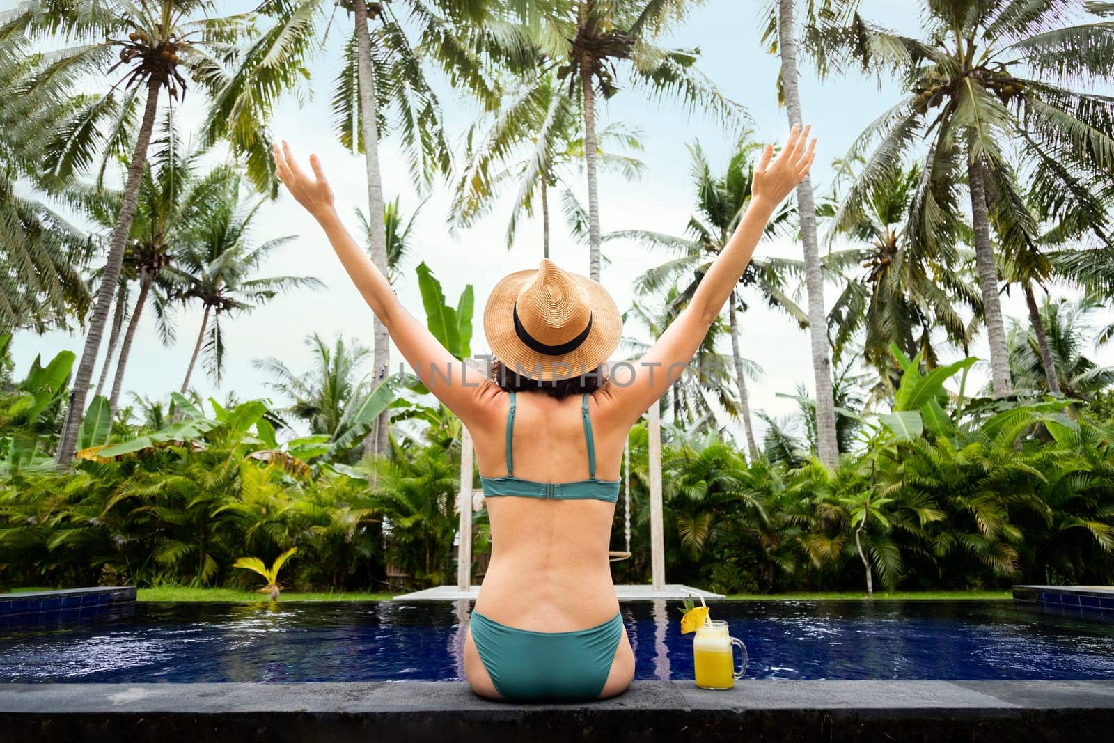 Back view of happy woman in bikini with arms up enjoying vacation on tropical paradise. Female relaxing in swimming pool, enjoying freedom in nature. Trip, vacation, mental health and healthy lifestyle concept.