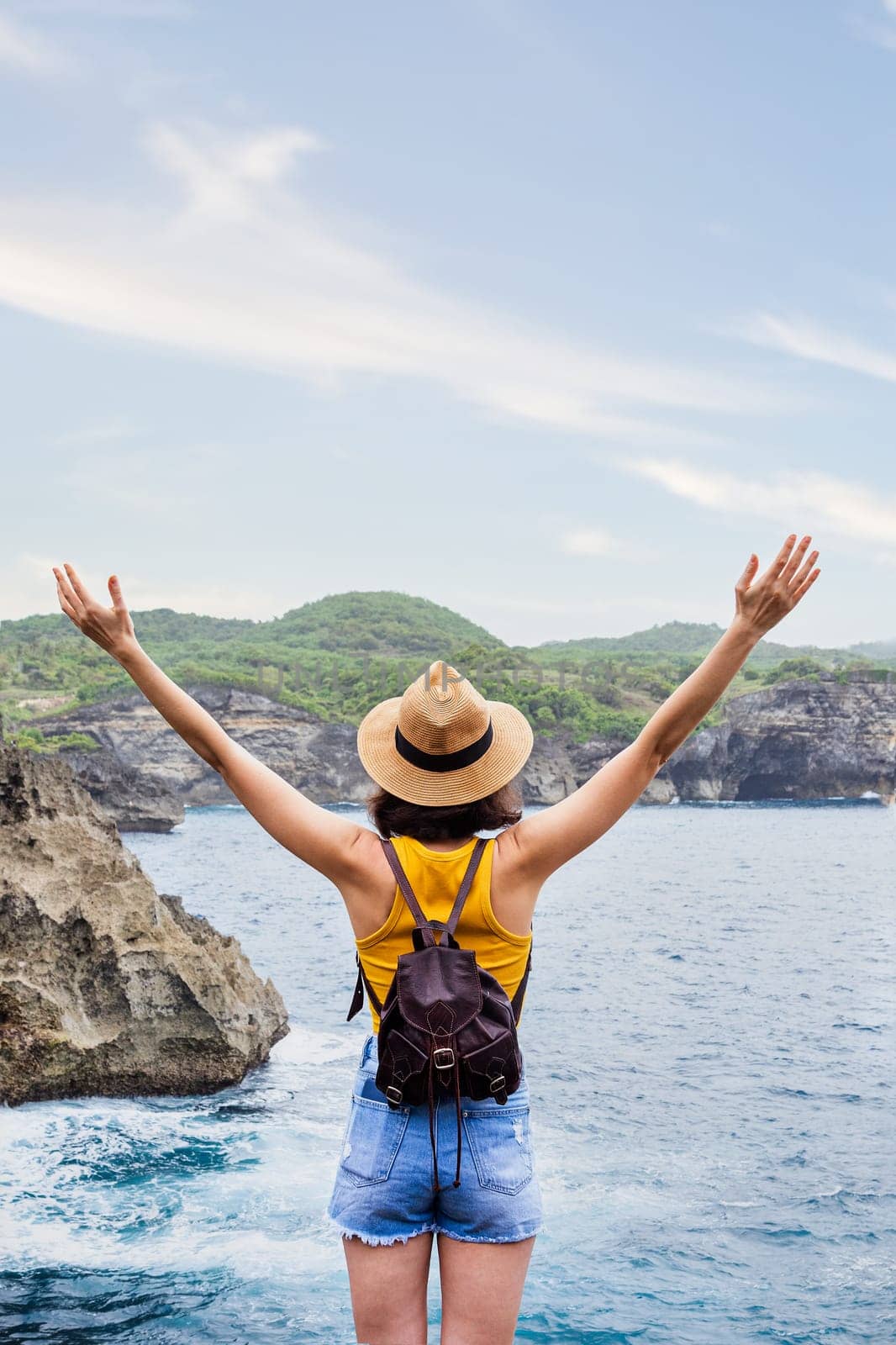 Back view of happy tourist woman with arms up enjoying vacation on Nusa Penida, Bali. Female enjoying freedom in nature. Vertical image. Copy space. Trip, vacation, mental health and healthy lifestyle concept.