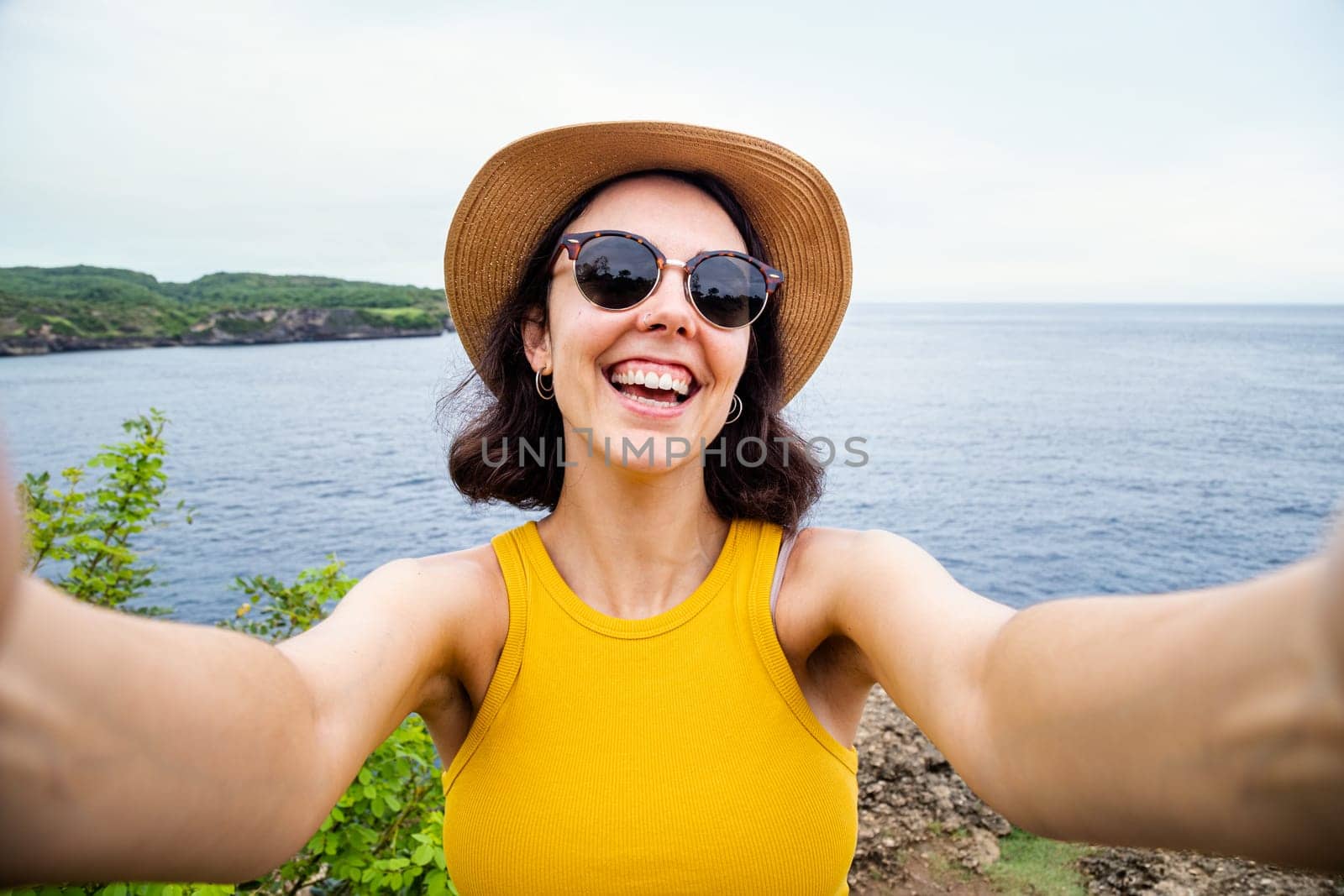 Happy young woman looking at camera taking selfie near the ocean during summer vacation. Travel, freedom and happiness concept.