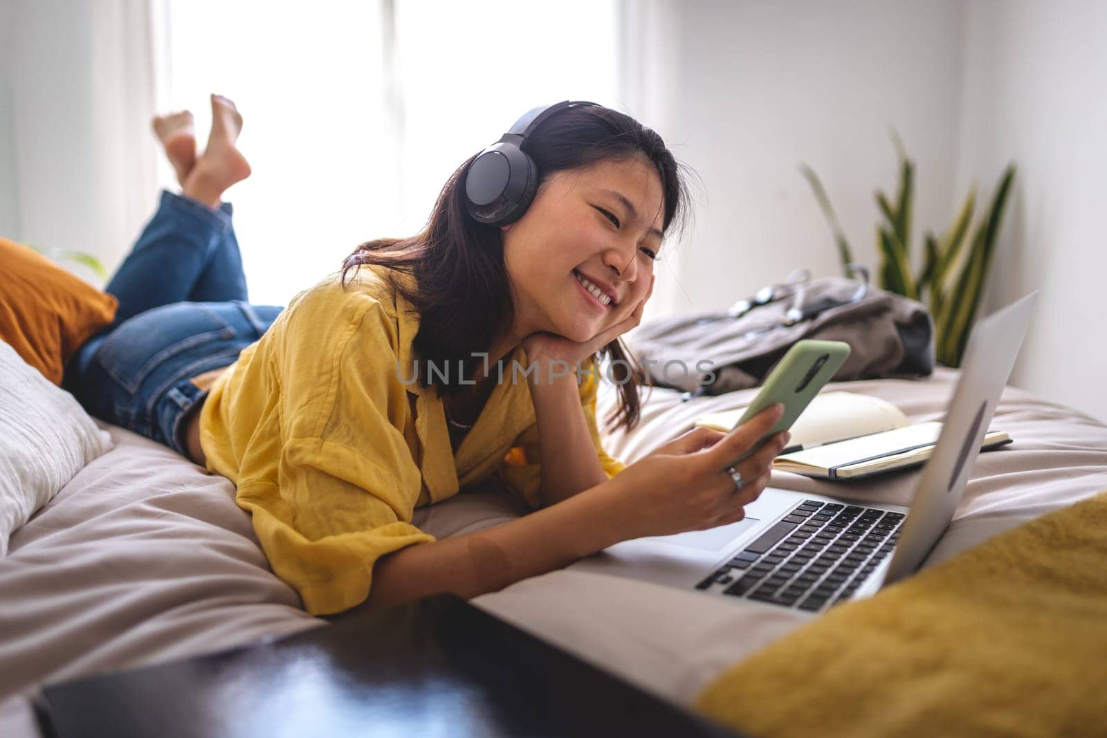 Female young chinese teen college student using phone and headphones listening to music lying down on bed. Asian young woman at home relaxing.