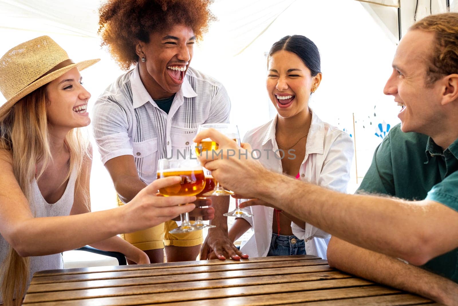 Young black man and his multiracial friends toasting with beer at a beach bar during summer vacation. by Hoverstock