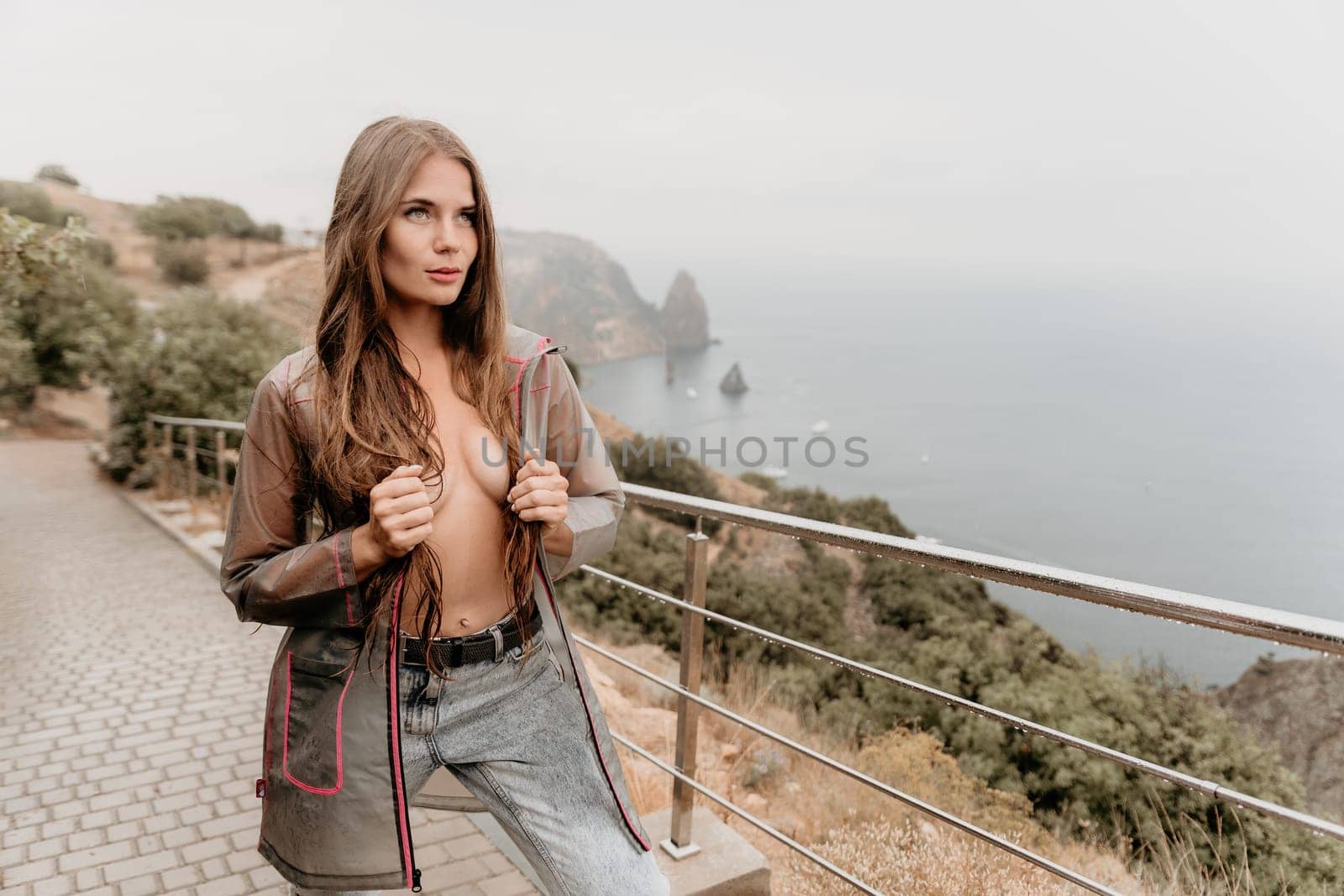 Woman rain park. Happy woman portrait wearing a raincoat with transparent umbrella outdoors on rainy day in park near sea. Girl on the nature on rainy overcast day