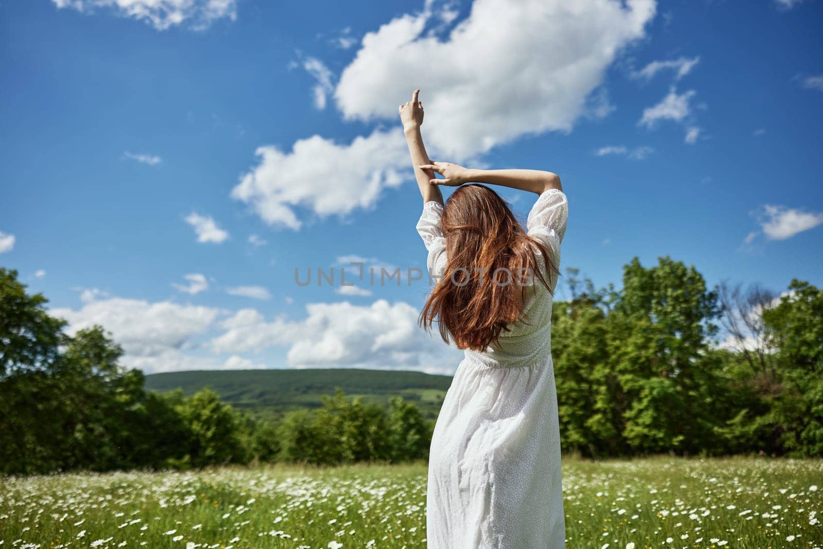 a woman stands with her back to the camera in a light dress in a chamomile field with her hands raised above her head. High quality photo