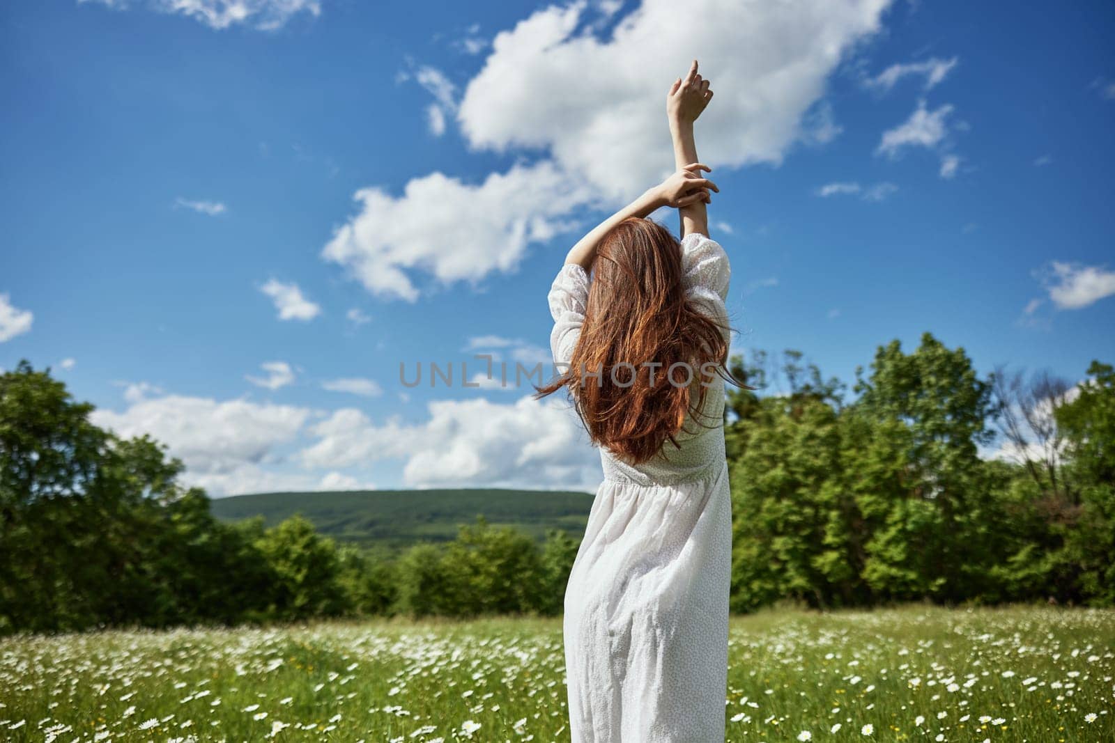 a woman stands with her back to the camera in a light dress in a chamomile field with her hands raised above her head. High quality photo