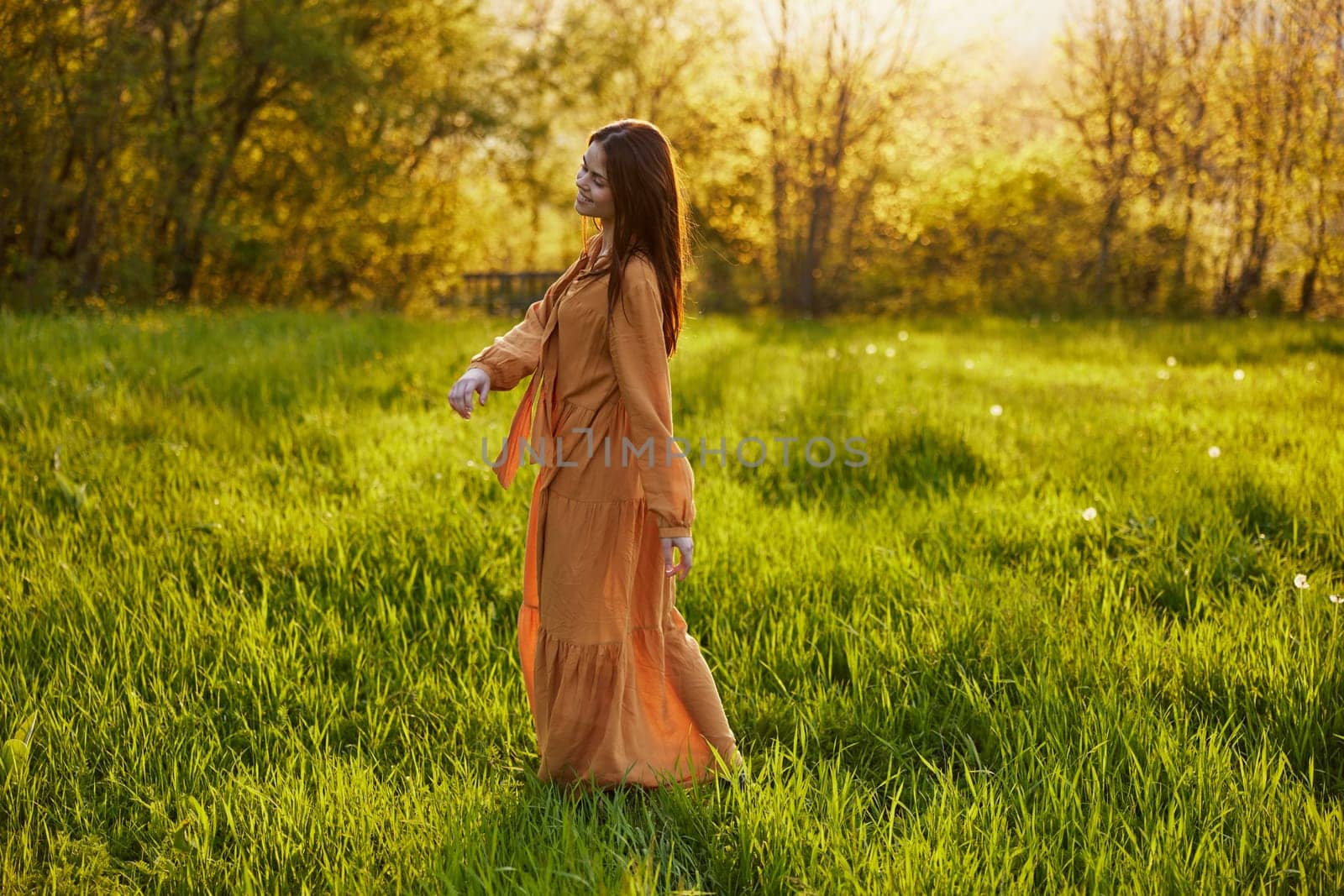 a relaxed slender woman enjoys the sunset standing in a green field with tall grass in an orange dress posing looking away by Vichizh