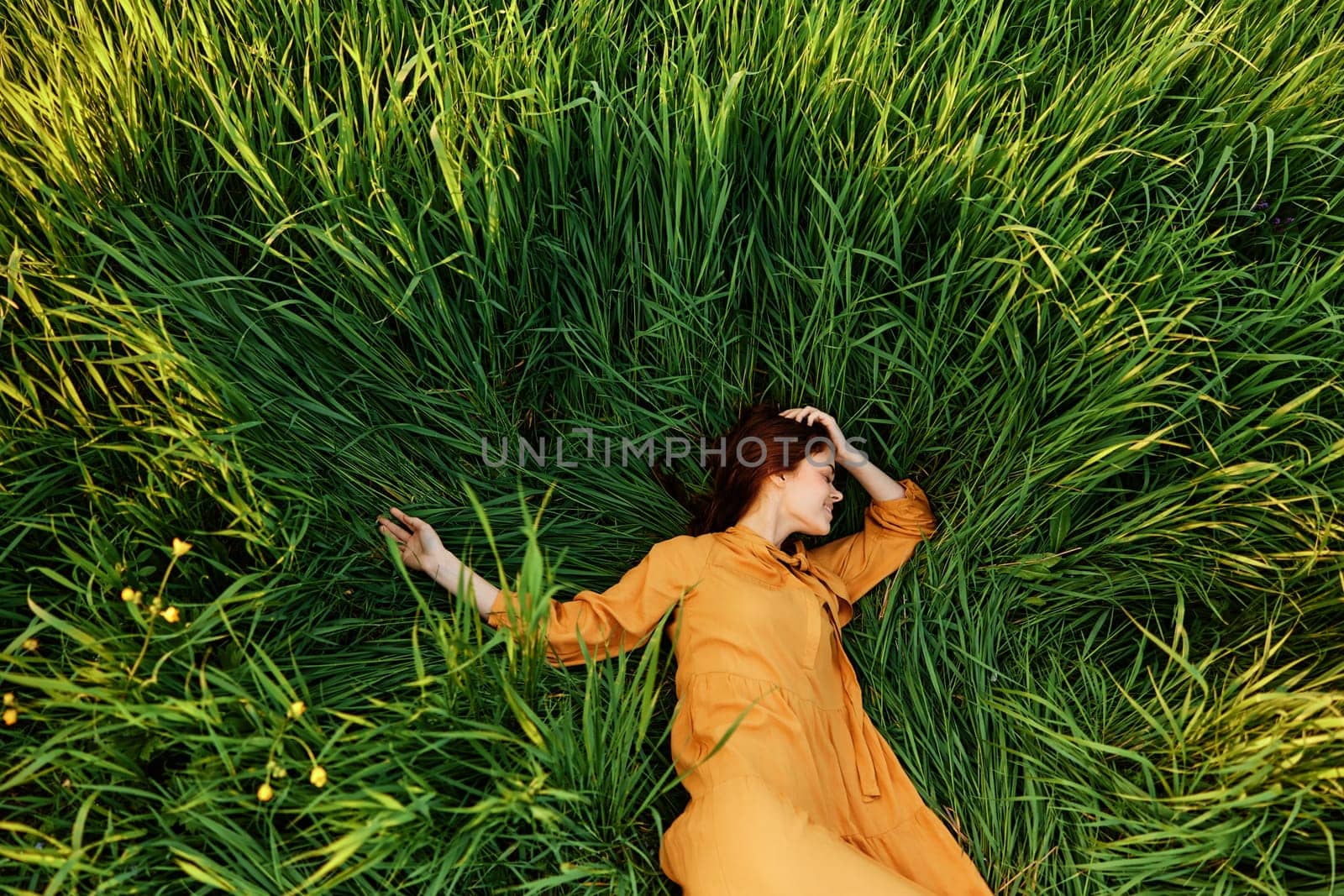 an elegant woman lies in the tall green grass in a long summer orange dress and with her arms outstretched enjoys nature and sunny weather with her eyes closed. High quality photo