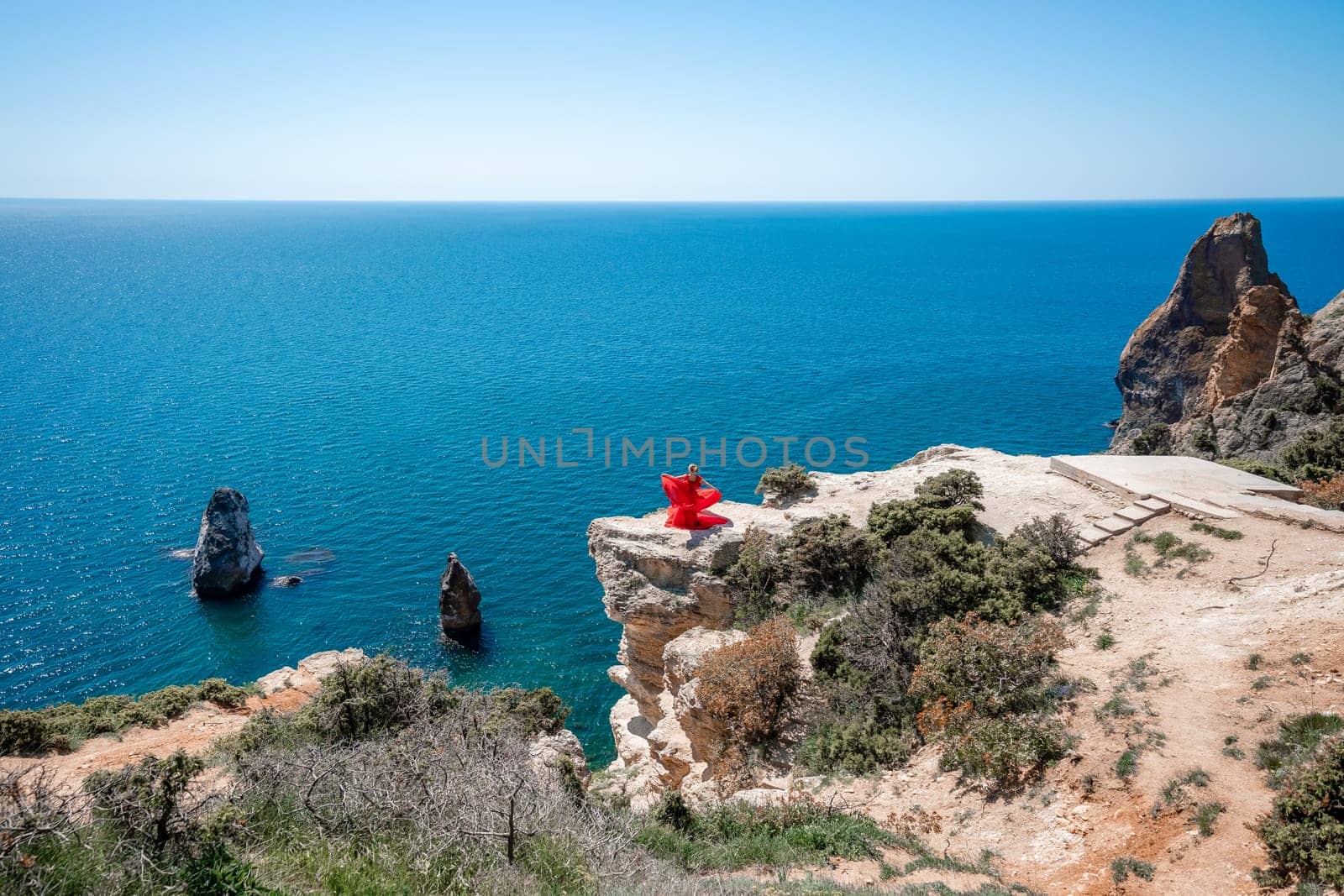 Woman in a red dress on the sea. Side view Young beautiful sensual woman in red long dress posing on a rock high above the sea at sunset. Girl in nature against the blue sky.