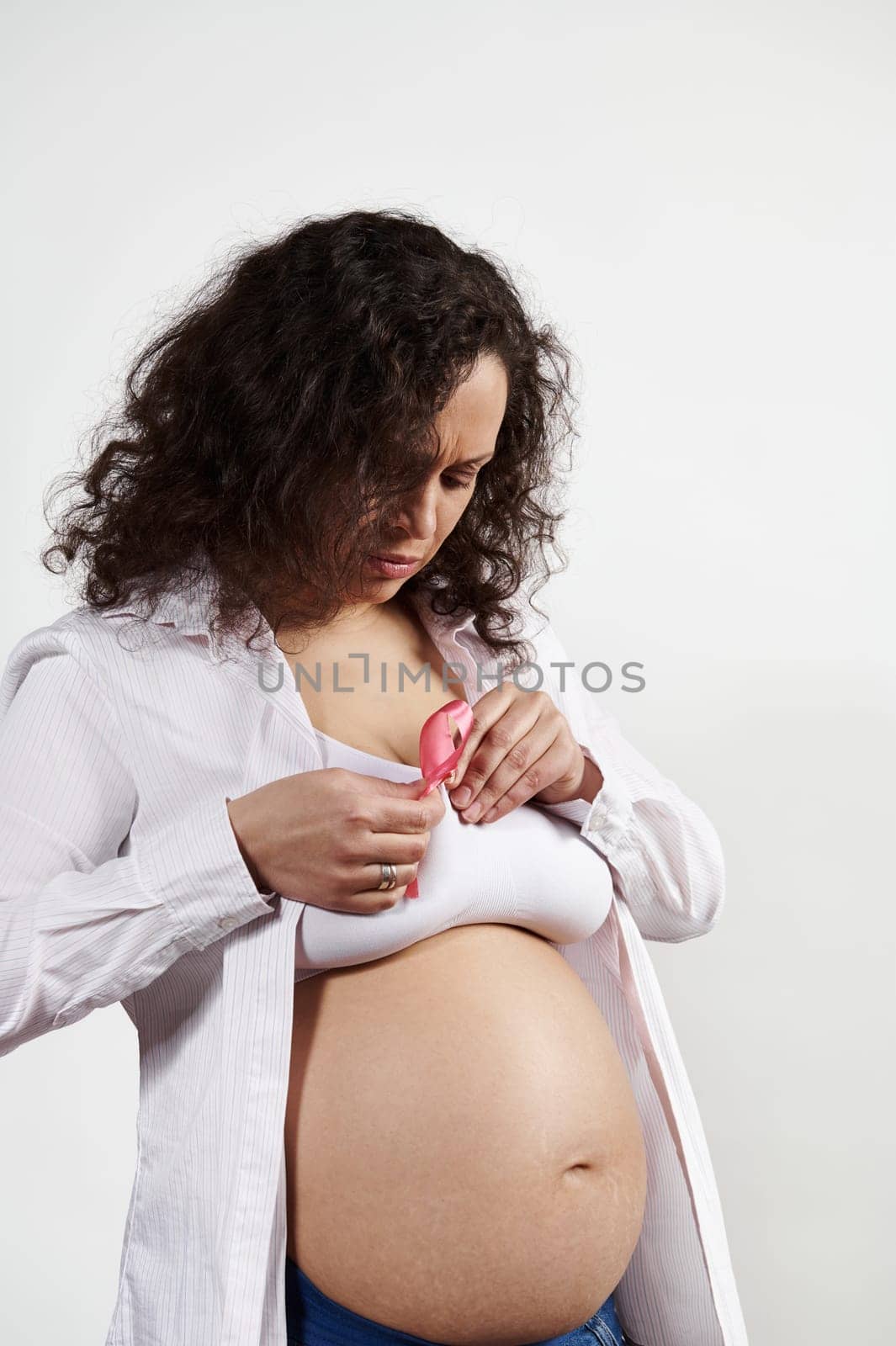 Multi ethnic pregnant woman puts pink satin ribbon on her bra, shows her support and compassion for all woman fighting breast cancer disease, posing bare belly isolated on white background. October 1