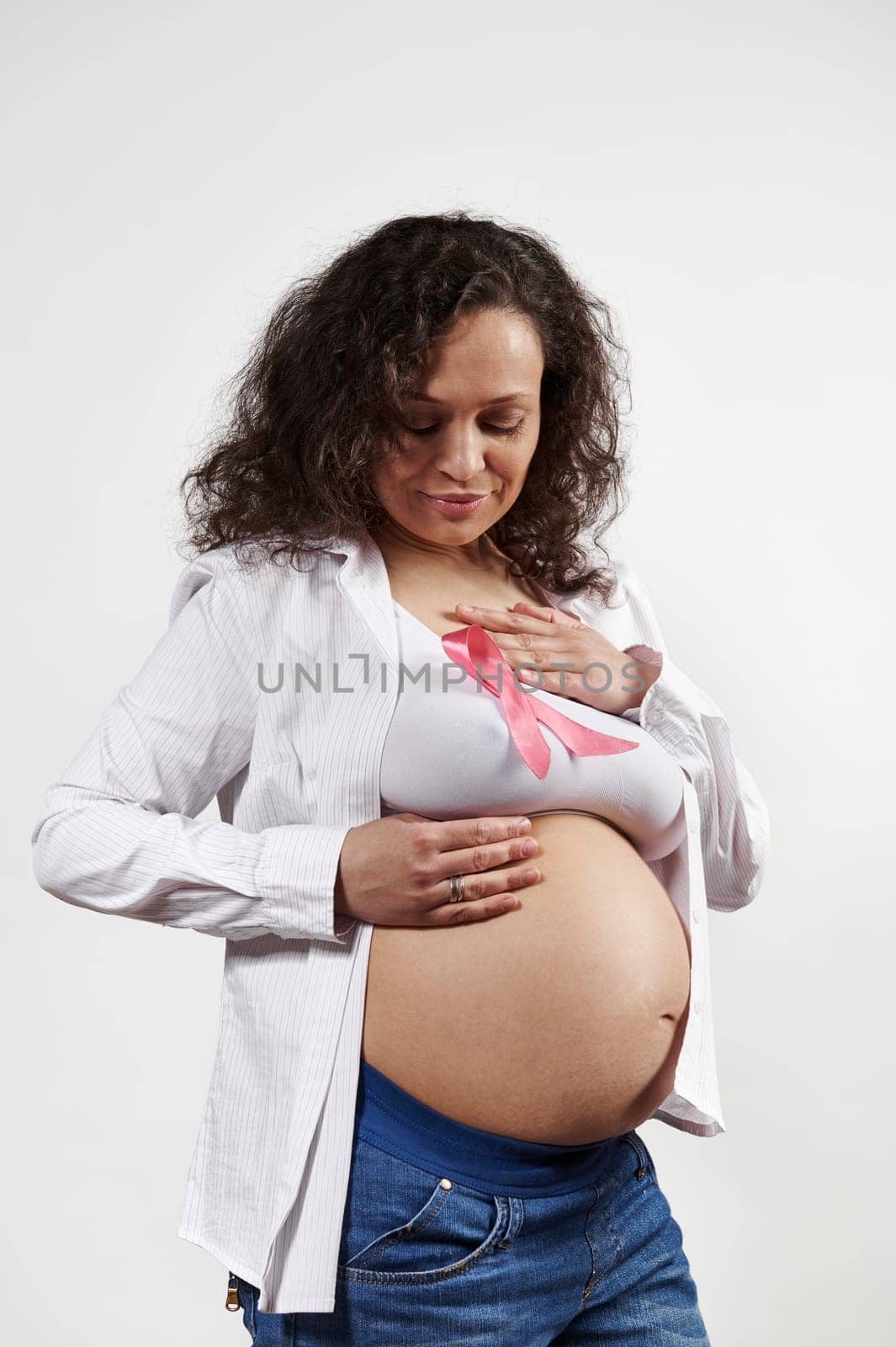 Smiling positive optimistic pregnant woman holding hands on a pink satin ribbon on her underwear, isolated on white background. Pregnancy. World Cancer Day. October 1st. Women's Health. Gynecology
