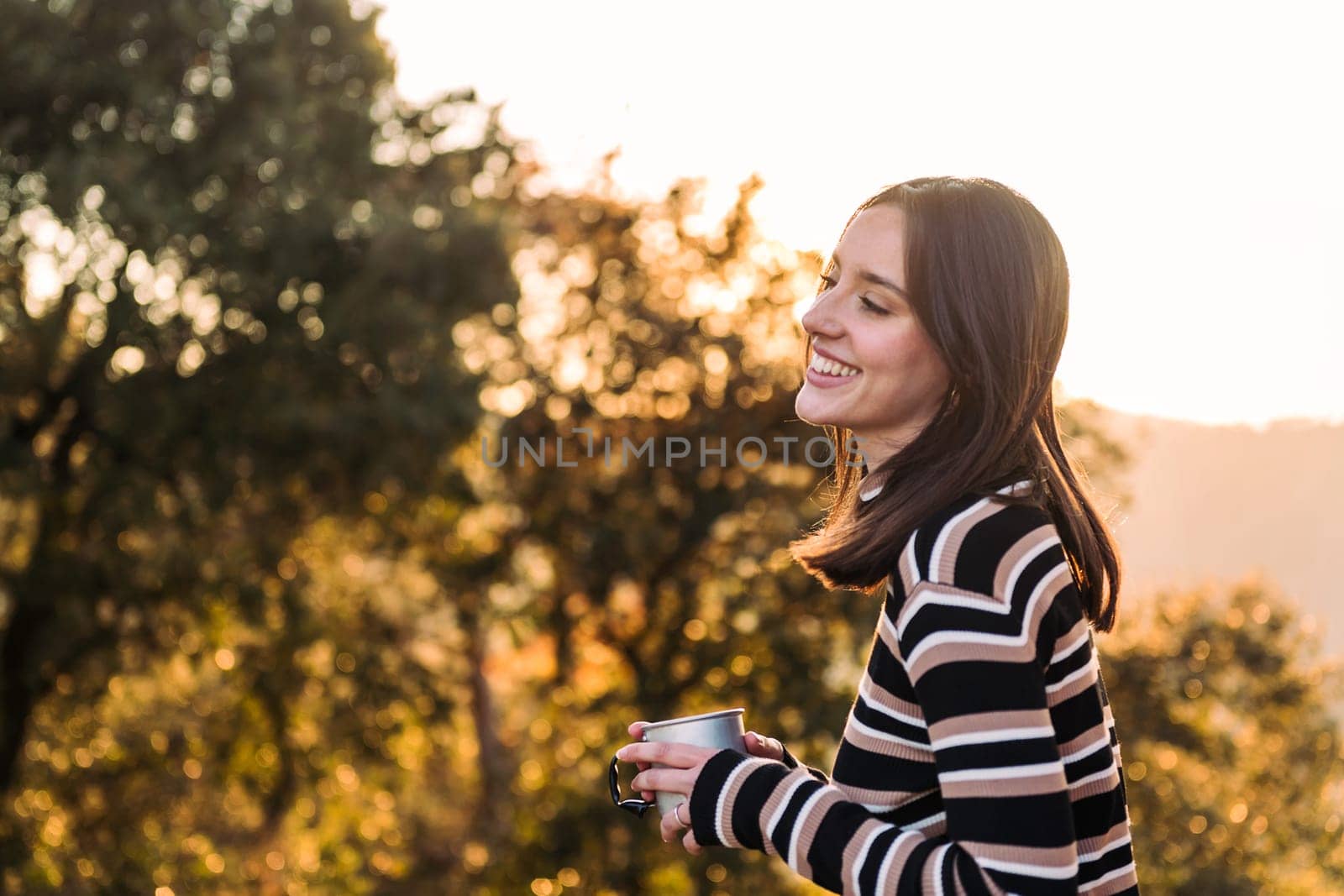 young woman smiling happy in nature with a mug of hot drink in hand, concept of morning bliss and wilderness lifestyle, copy space for text