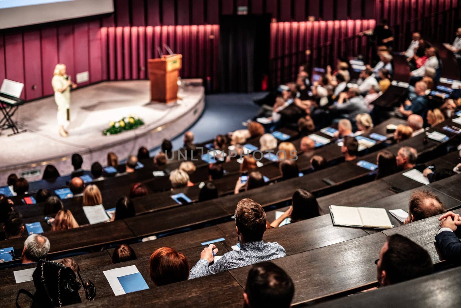 Business and entrepreneurship symposium. Female speaker giving a talk at business meeting. Audience in conference hall. Rear view of unrecognized participant in audience.