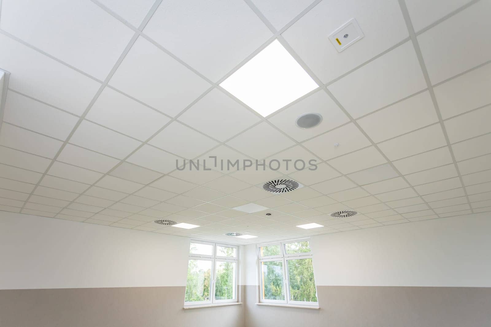 Suspended panel ceiling seen from below with lamps lit. Just below the ceiling are two windows.