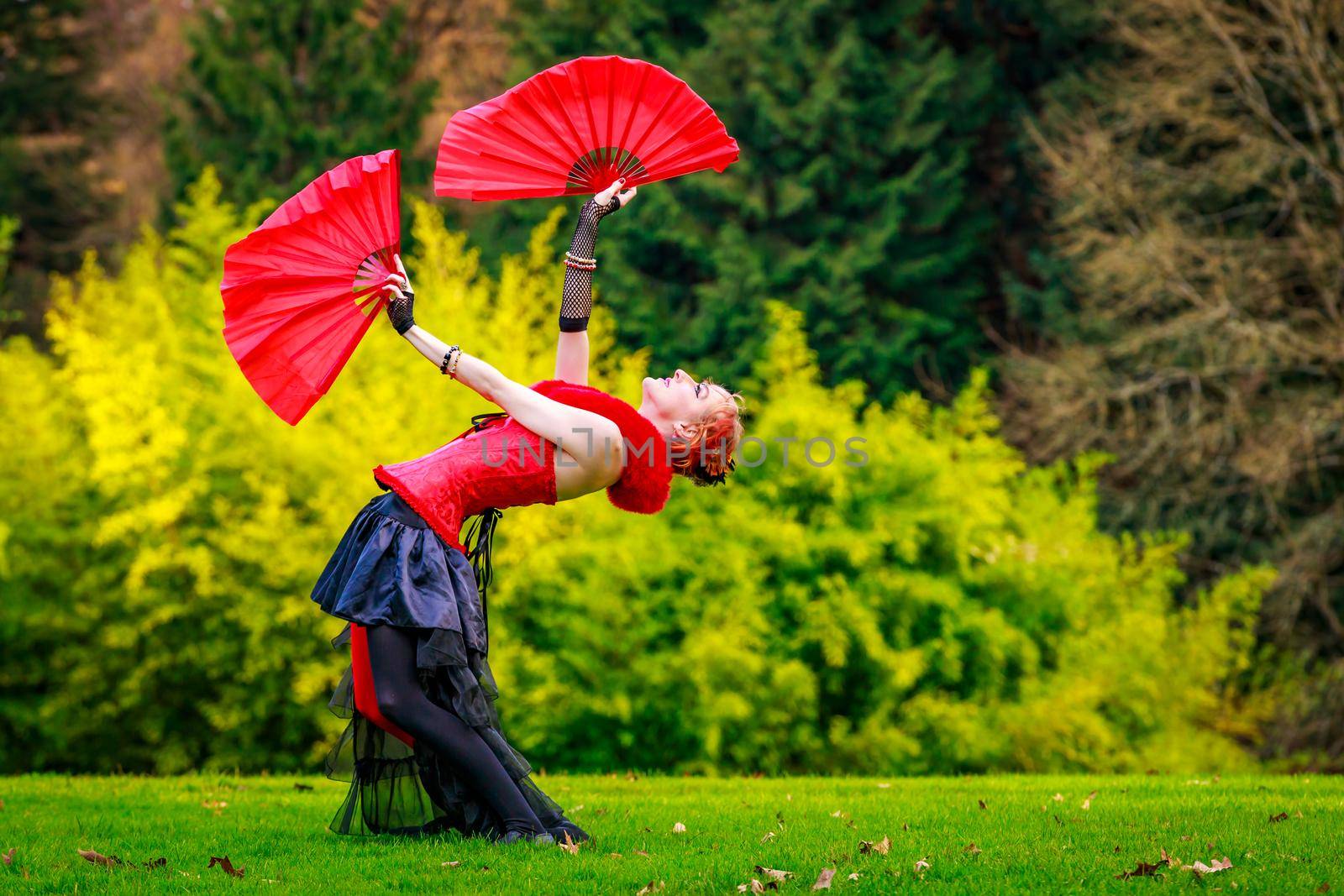 Young beautiful woman in circus costume play with red fans in the park.