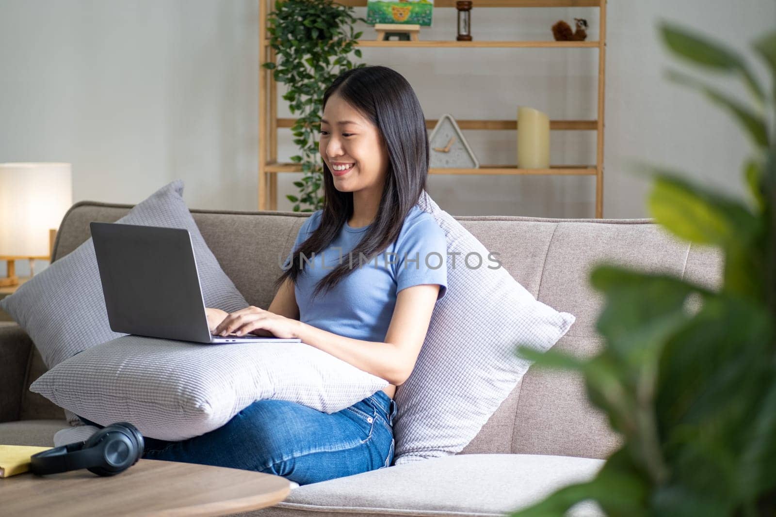 Happy cheerful young asian woman sitting on sofa with laptop, smart beautiful young asian woman chilling in house with by wuttichaicci