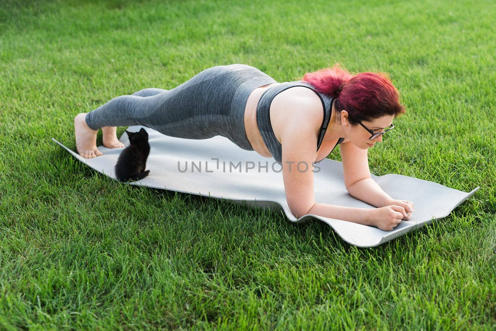 Young plus size woman in sporty top and leggings standing in plank on yoga mat spending time on green grass in yard. Black kitten walks around her. Well being and body positivity fitness concept by Satura86
