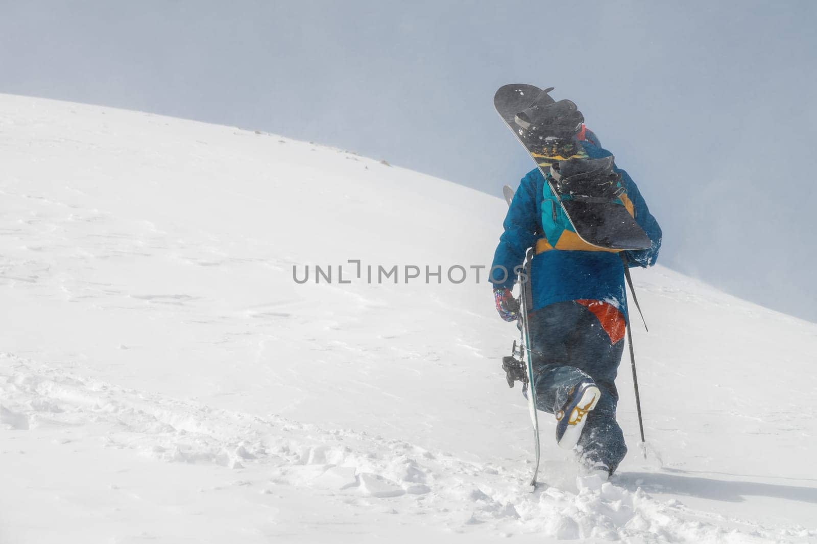 man with ski equipment and a snowboard climbs a snowy mountain. Blue sky and snowy mountain in the background. by yanik88