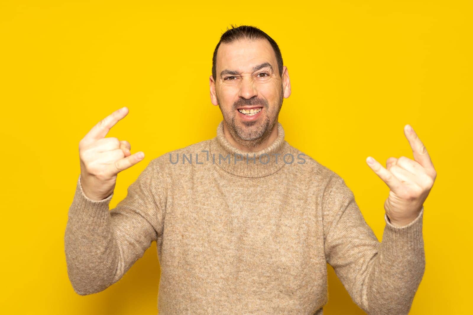 Photo of excited funky guy dressed in beige turtleneck sweater showing two heavy rock gestures over isolated yellow color background