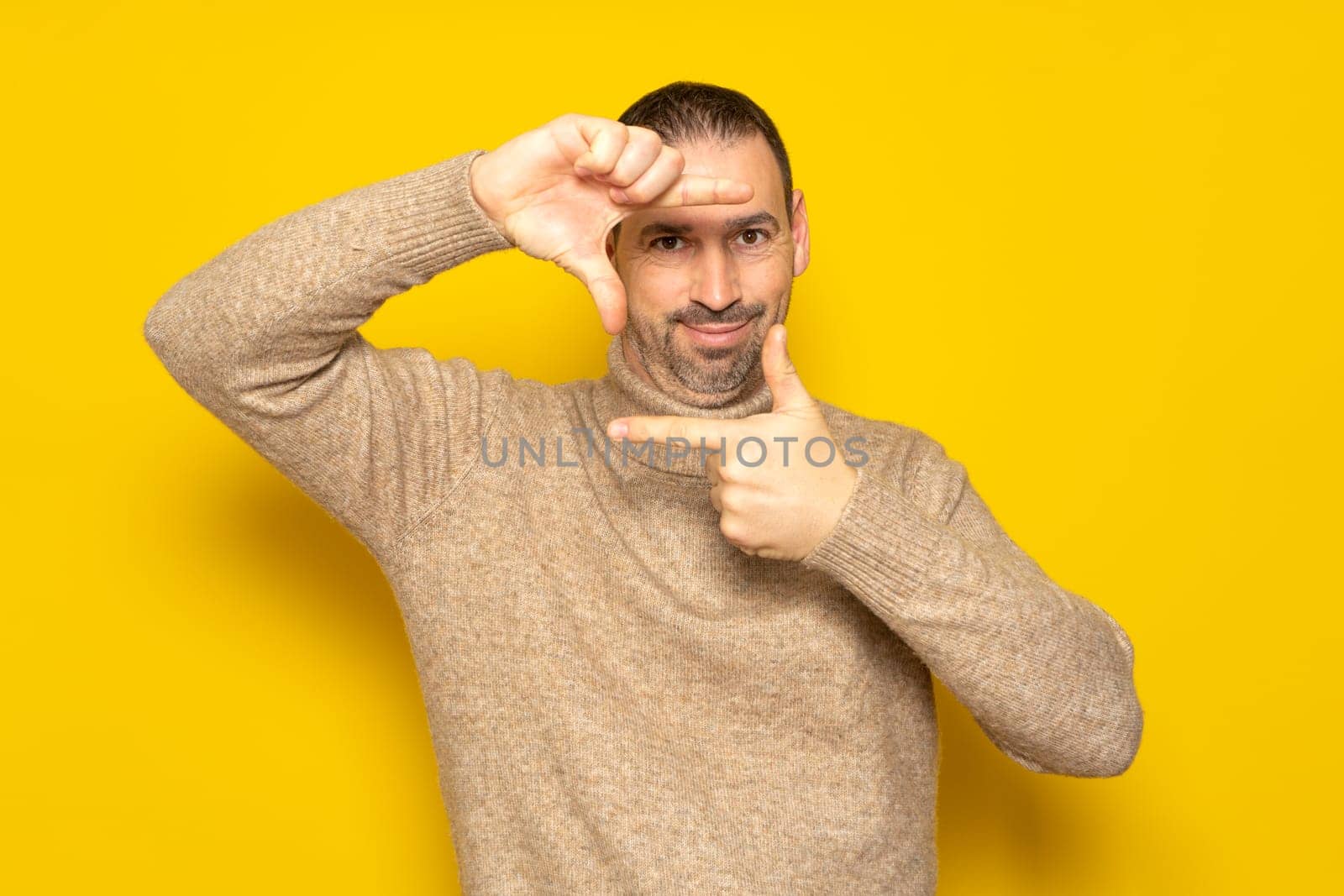 Handsome bearded happy male blink with eye and looking through a frame formed by his hands. Attractive man make frame by fingers, isolated on yellow background with copy space for text.