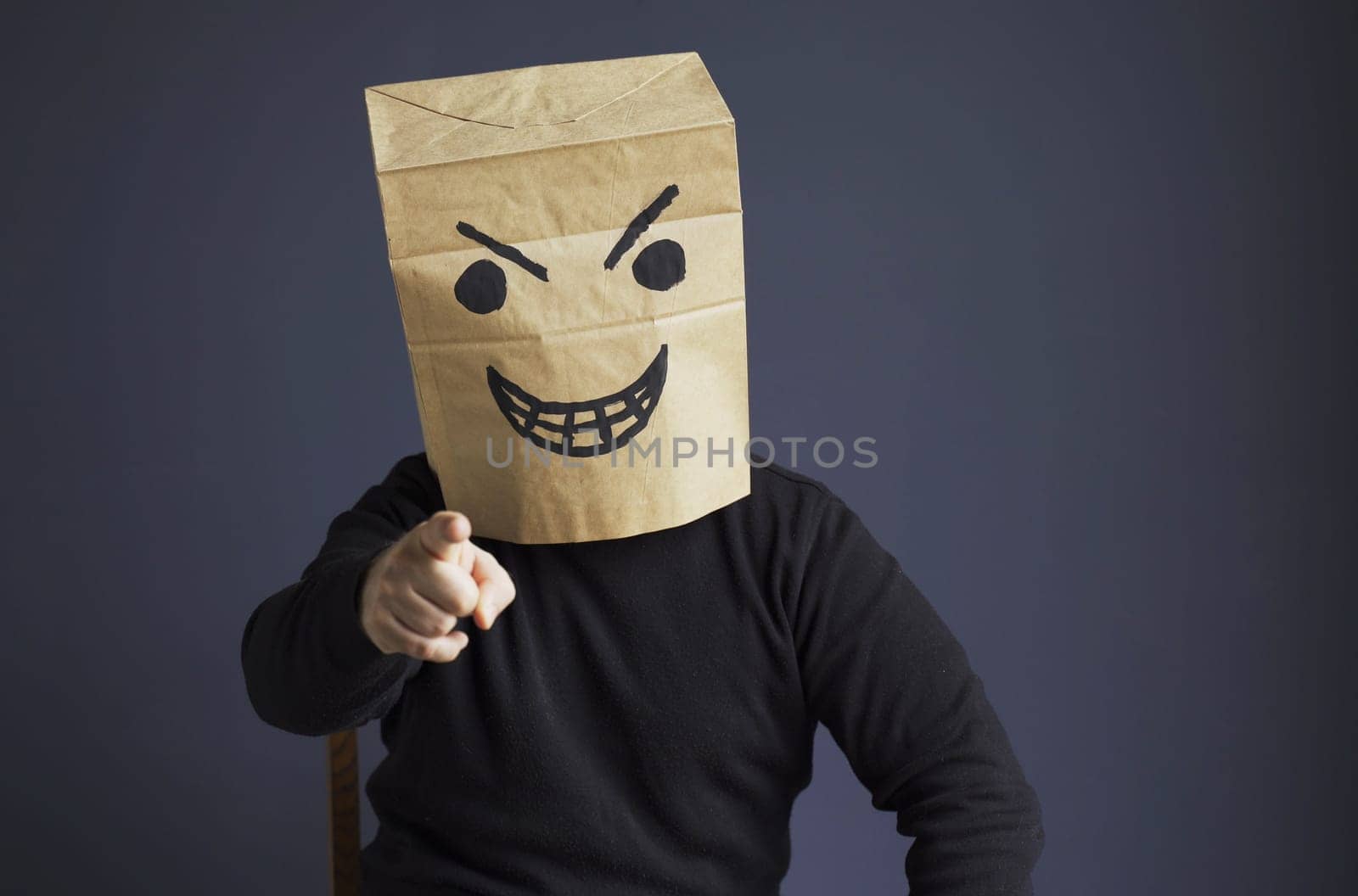 A man in a black turtleneck with a paper bag with an angry emoticon on his head shows his hand pointing forward. Emotions and anger
