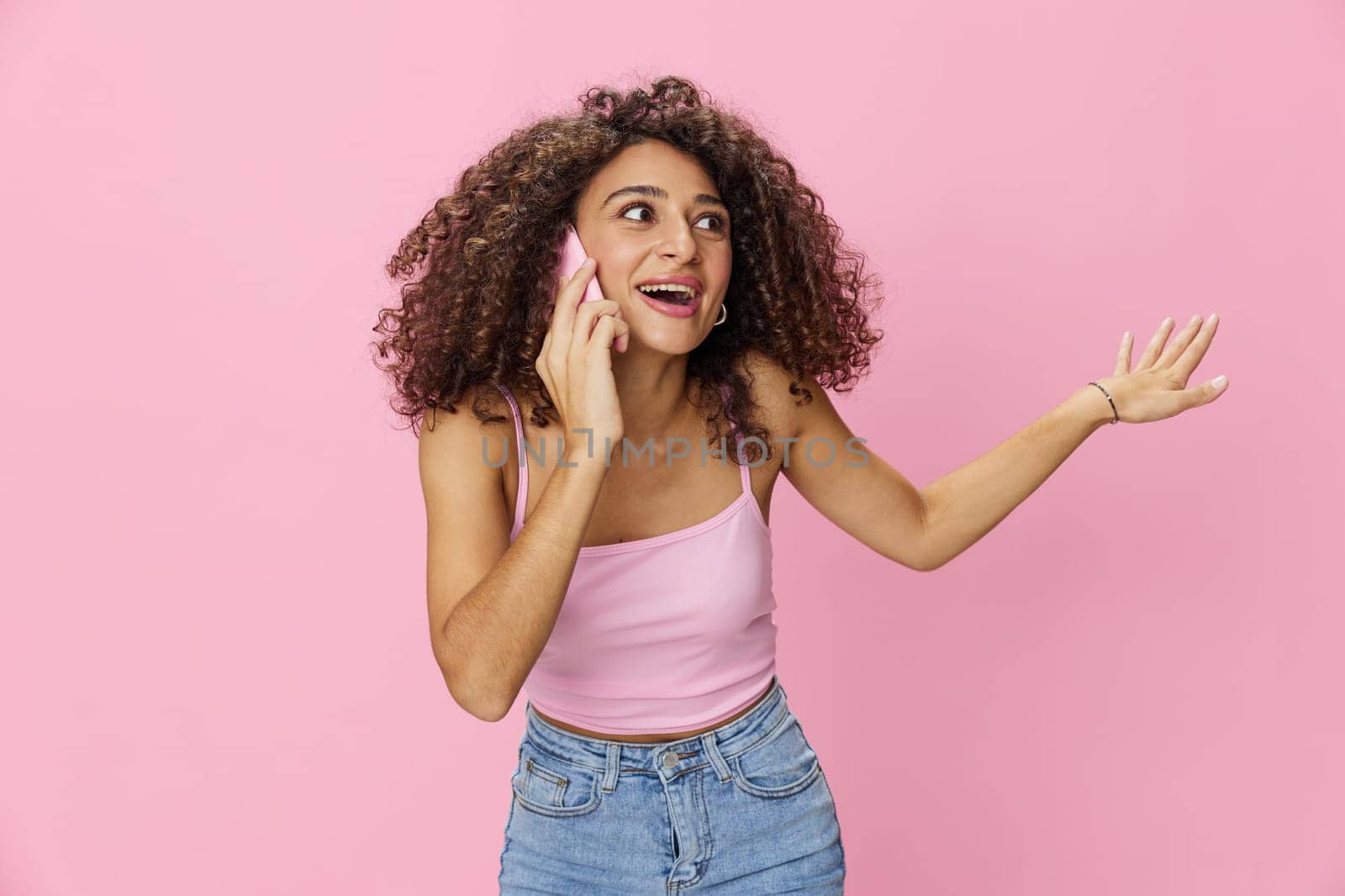 Woman with curly afro hair talking on the phone in a pink top and jeans on a pink background, smile, happiness, finger pointing copy space. High quality photo