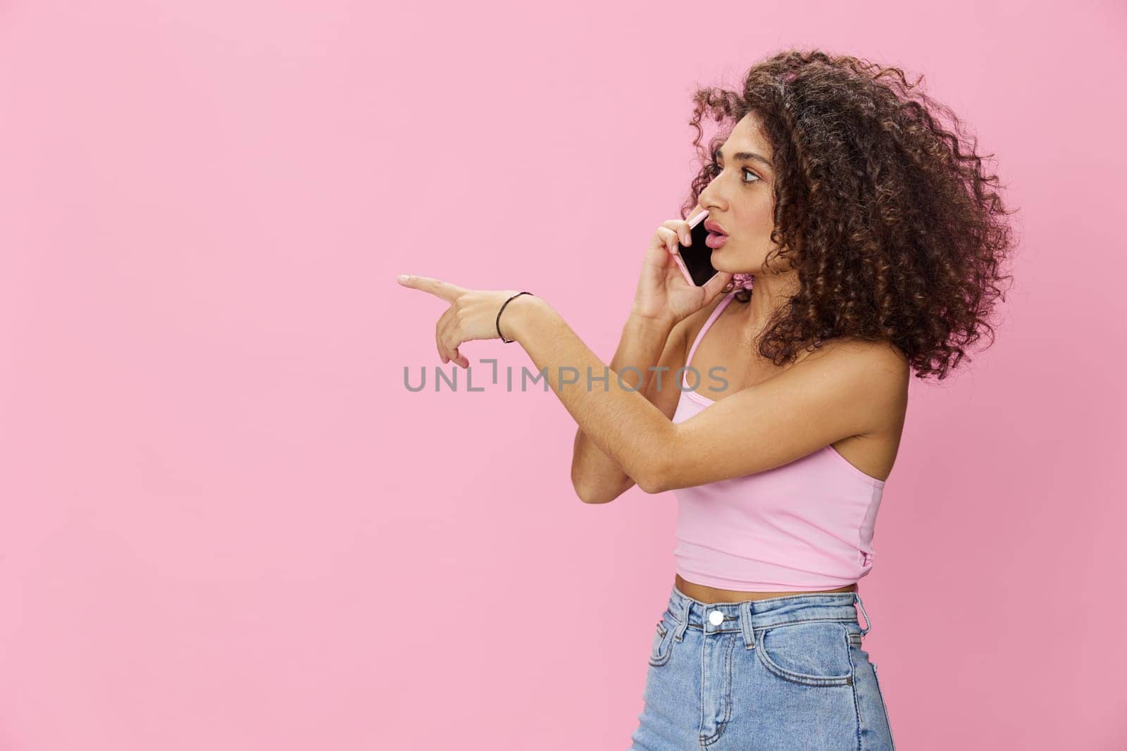 Woman with curly afro hair talking on the phone in a pink top and jeans on a pink background, smile, happiness, finger pointing copy space. High quality photo