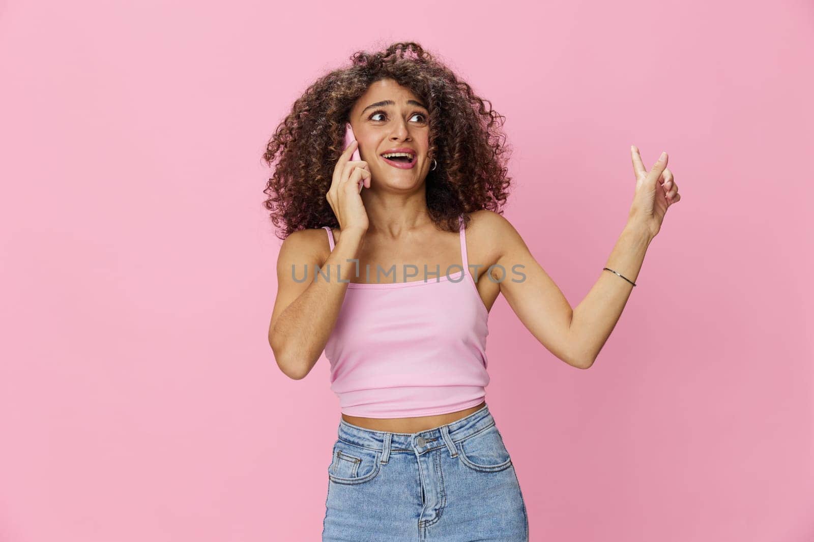 Woman with curly afro hair talking on the phone in a pink top and jeans on a pink background, smile, happiness, finger pointing copy space. High quality photo