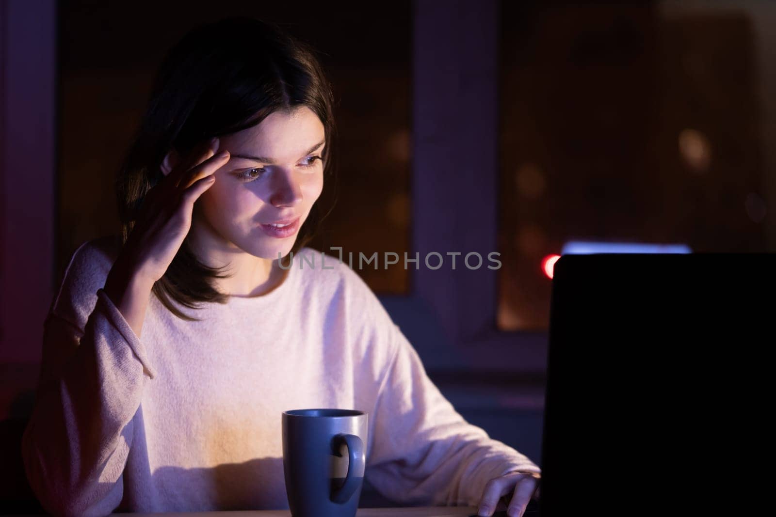 Young girl works at a laptop online at night against the background with the city lights outside the window. Woman is drinking coffee, holding a cup in her hands and reading the text on her device.