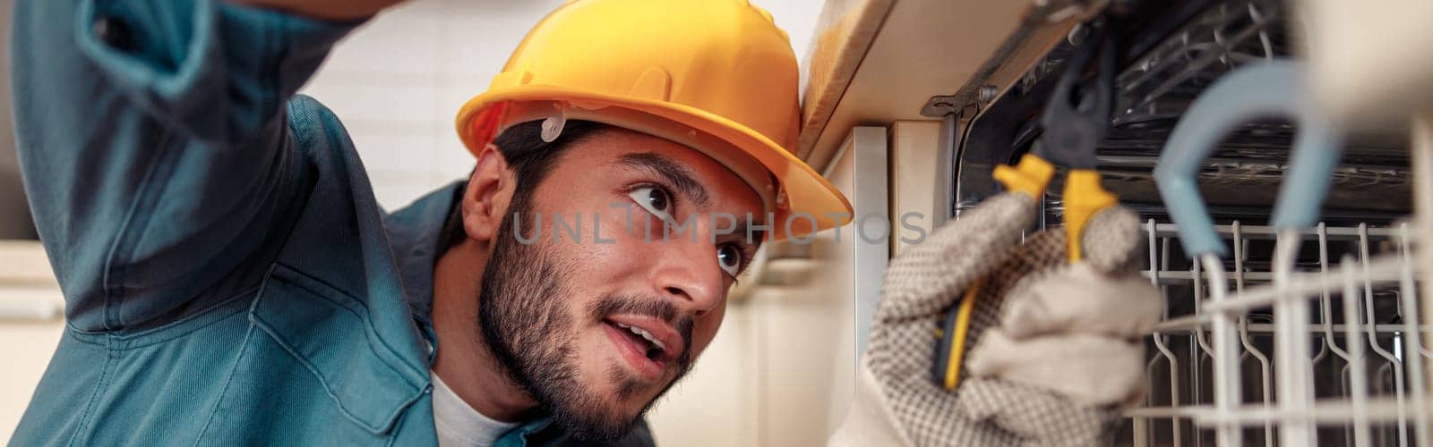 Close up of handyman in special clothing repairing dishwasher in modern kitchen by Yaroslav_astakhov