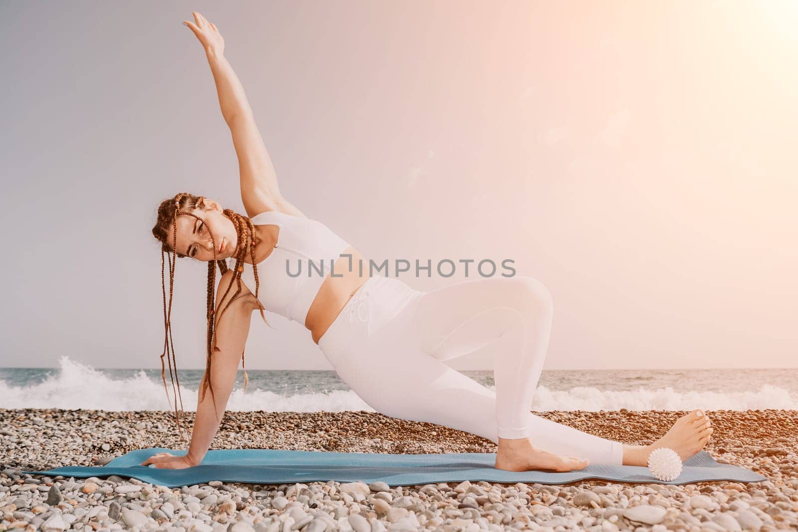 Woman yoga sea. Well looking middle aged woman with braids dreadlocks in white leggings and tops doing stretching pilates on yoga mat near sea. Female fitness yoga routine concept. Healthy lifestyle