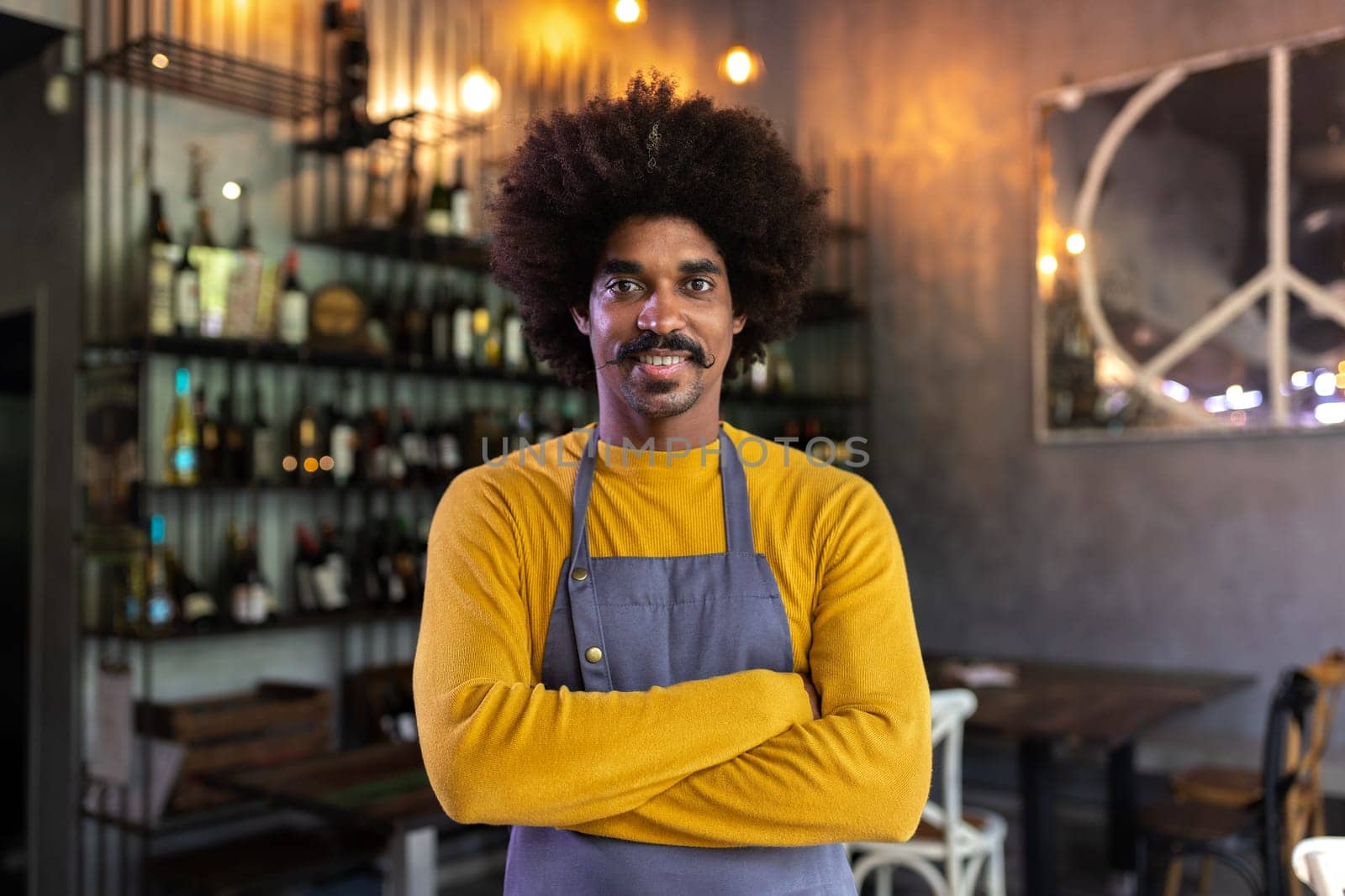 Happy African American waiter wearing apron standing in bar with arms crossed looking at camera. Restaurant concept.