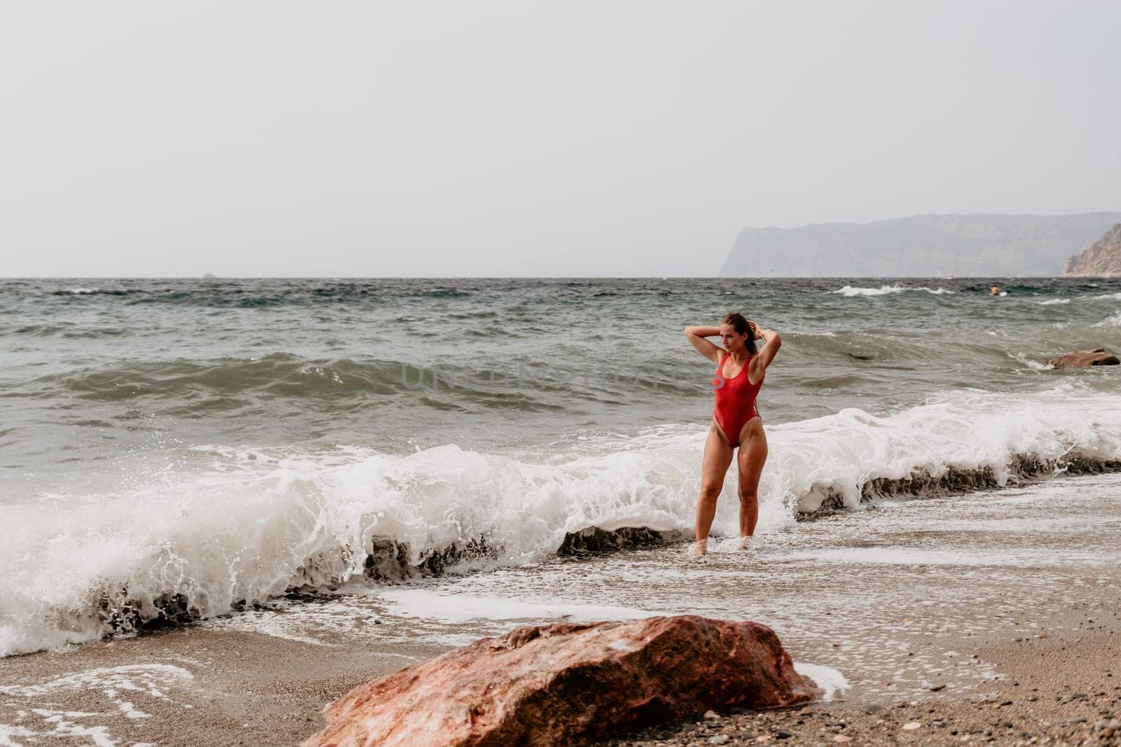 Woman travel sea. Young Happy woman in a long red dress posing on a beach near the sea on background of volcanic rocks, like in Iceland, sharing travel adventure journey