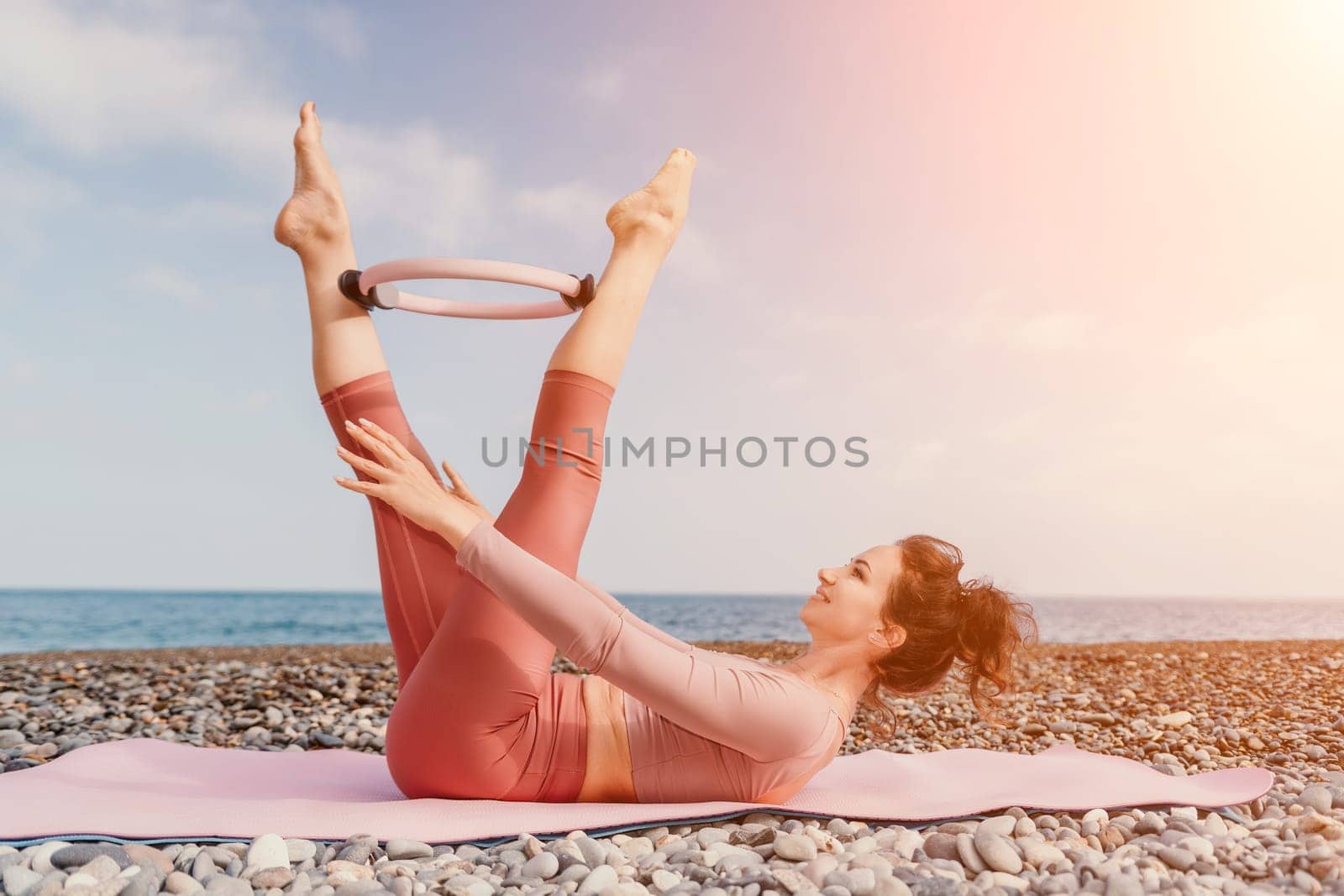 Middle aged well looking woman with black hair doing Pilates with the ring on the yoga mat near the sea on the pebble beach. Female fitness yoga concept. Healthy lifestyle, harmony and meditation.