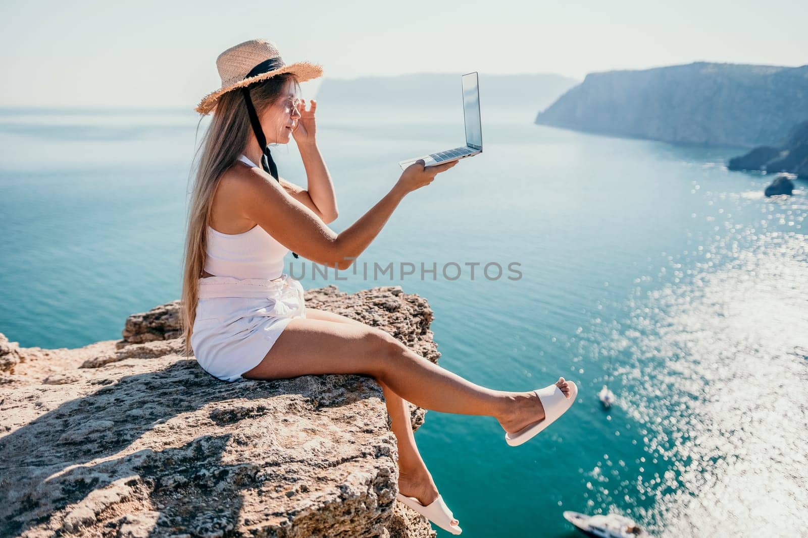 Working remotely on seashore. Happy successful woman female freelancer in straw hat working on laptop by the sea at sunset. Freelance, remote work on vacation