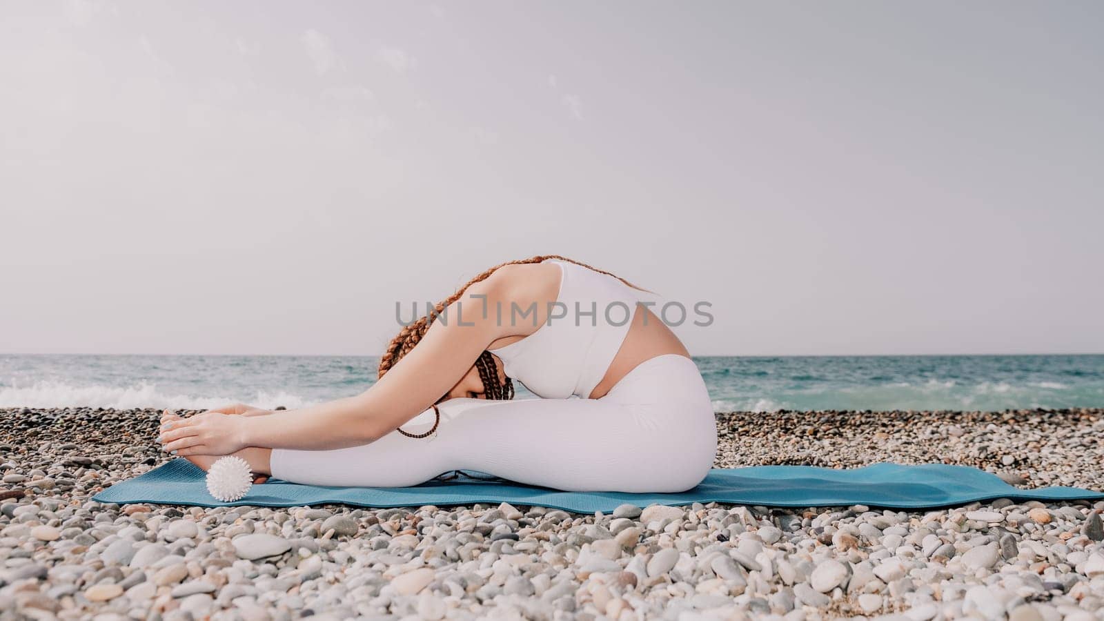 Woman yoga sea. Well looking middle aged woman with braids dreadlocks in white leggings and tops doing stretching pilates on yoga mat near sea. Female fitness yoga routine concept. Healthy lifestyle