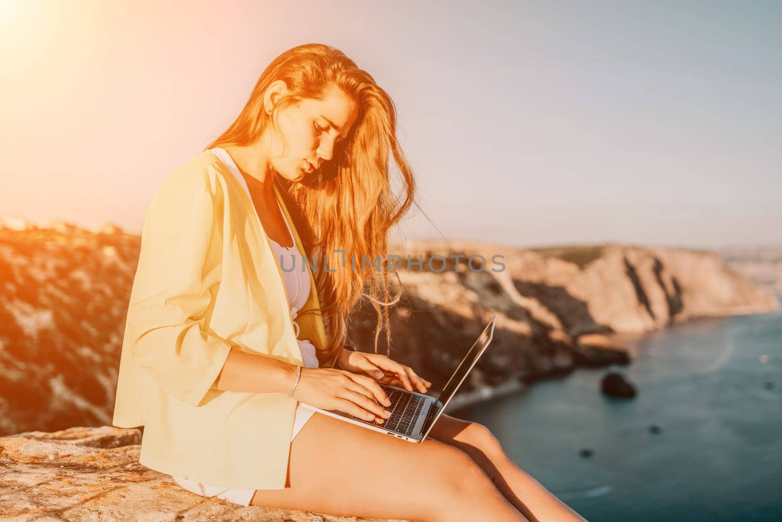 Successful business woman in yellow hat working on laptop by the sea. Pretty lady typing on computer at summer day outdoors. Freelance, travel and holidays concept.