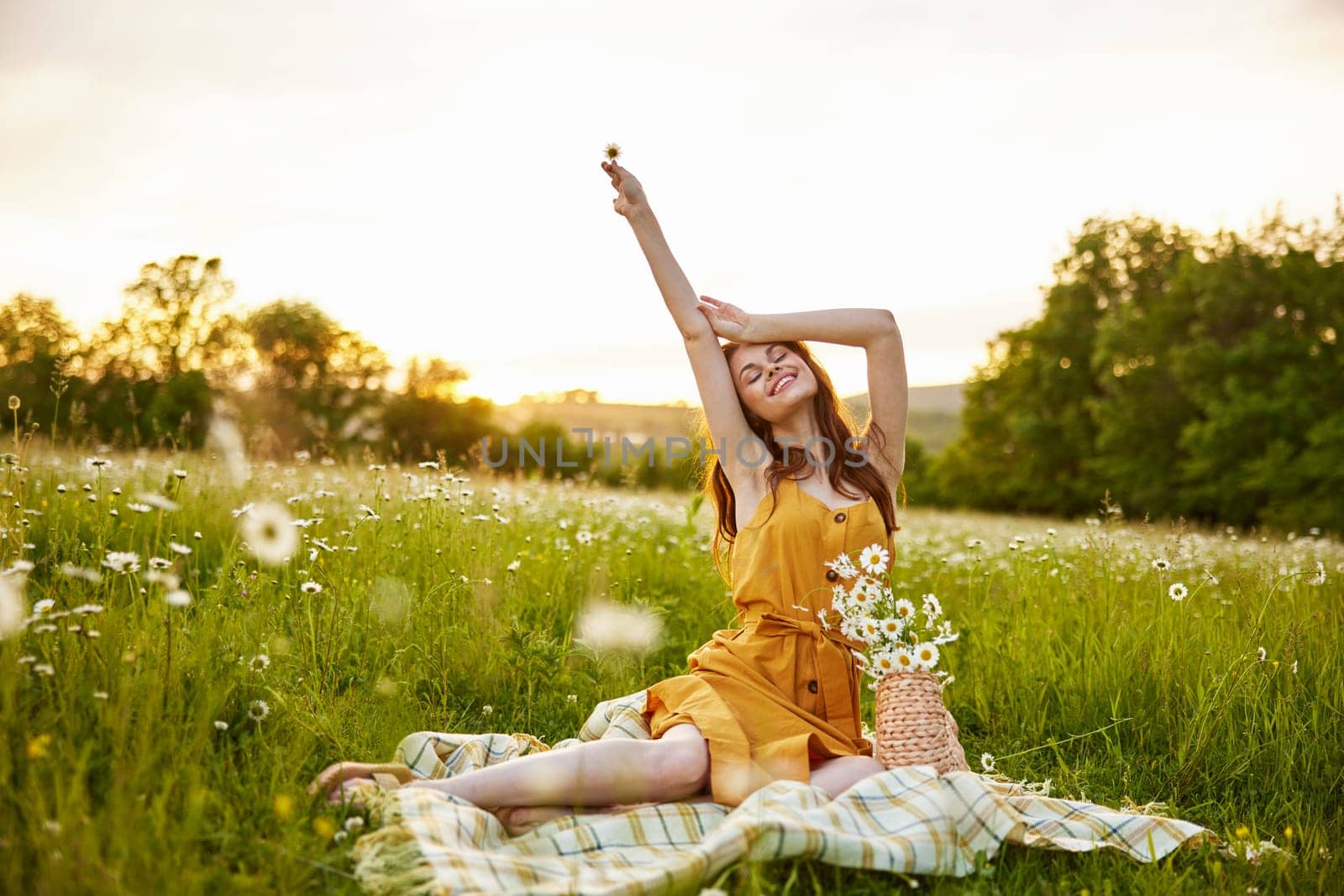a woman in an orange dress sits on a plaid in a chamomile field and happily raises her hands up. High quality photo
