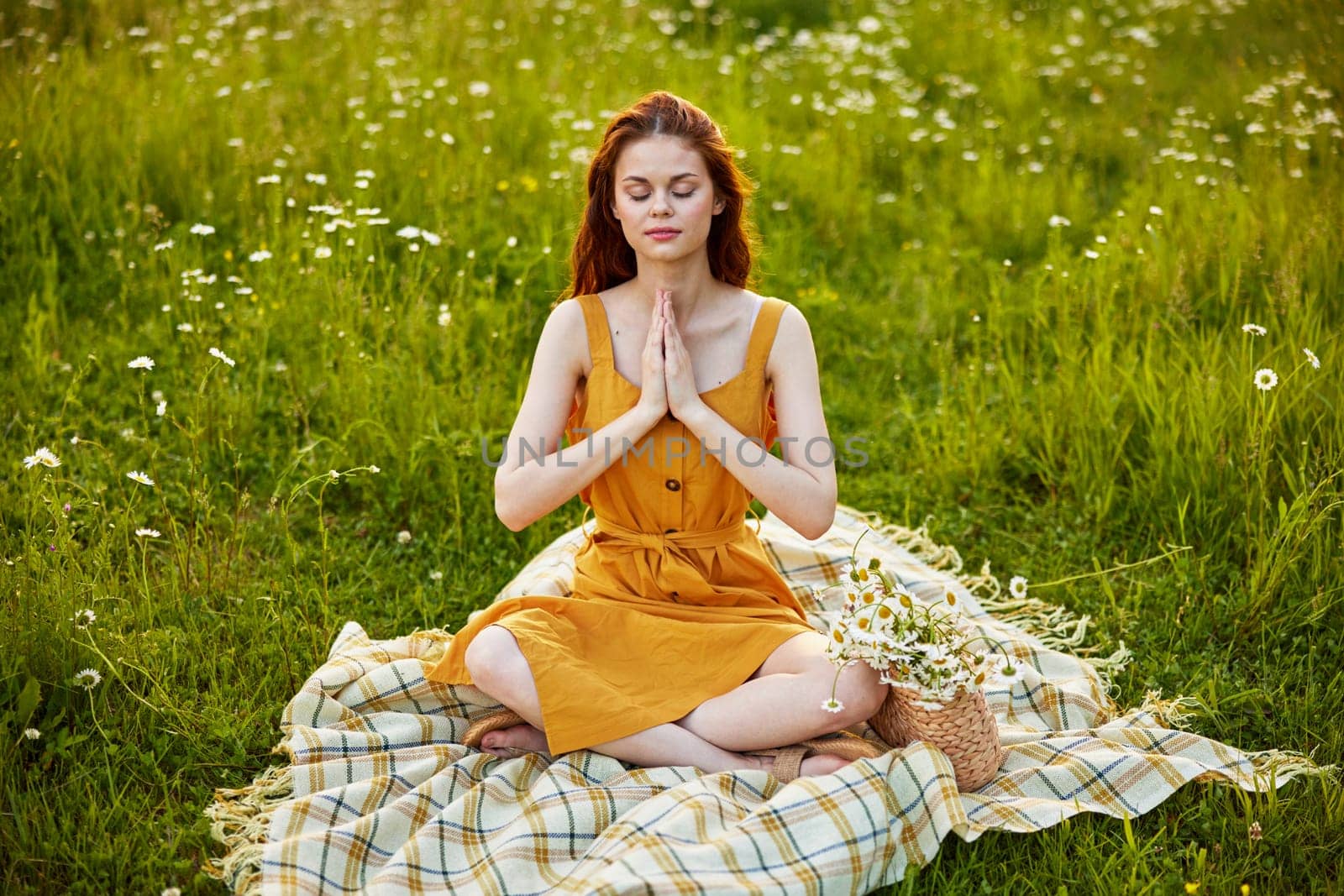 a woman in a chamomile field meditates holding her hands on her chest while sitting on a plaid by Vichizh