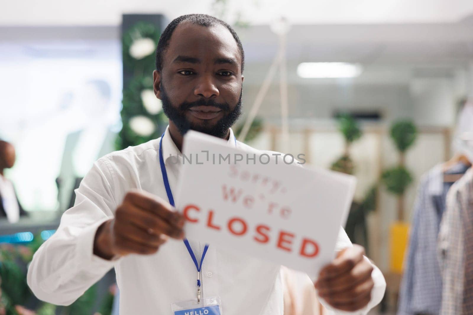 Fashion garment department store african american employee hanging closed sign on front door. Apparel shop worker holding signboard informing customers about work day end