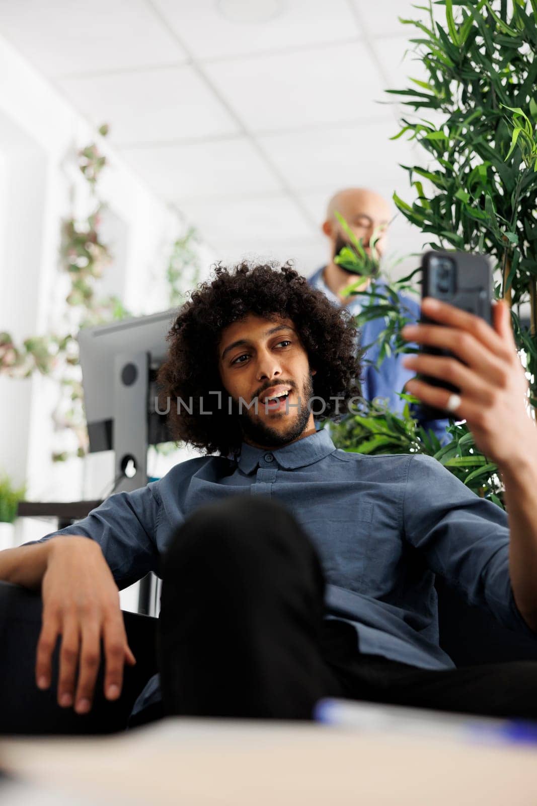 Young arab man having virtual video communication on smartphone in corporate office. Smiling start up company employee chatting online with coworkers team using mobile phone