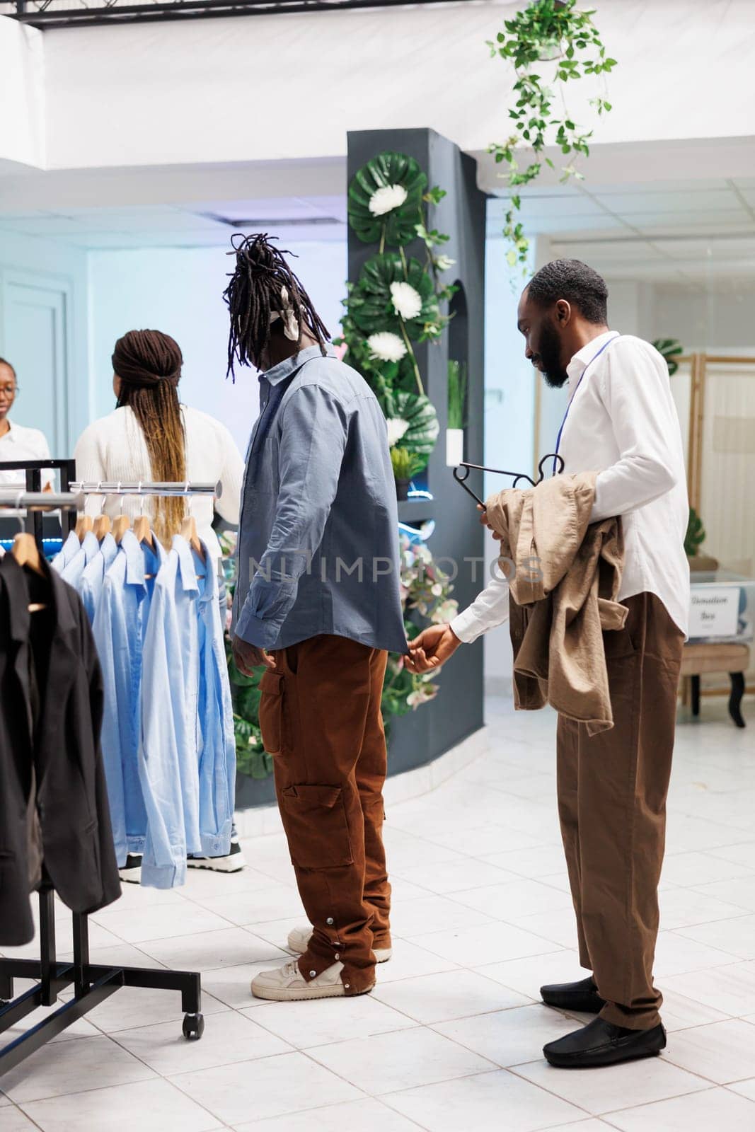 Clothing store employee examining shirt on customer and checking fit. African american man choosing menswear in shopping mall while getting help from fashion boutique assistant