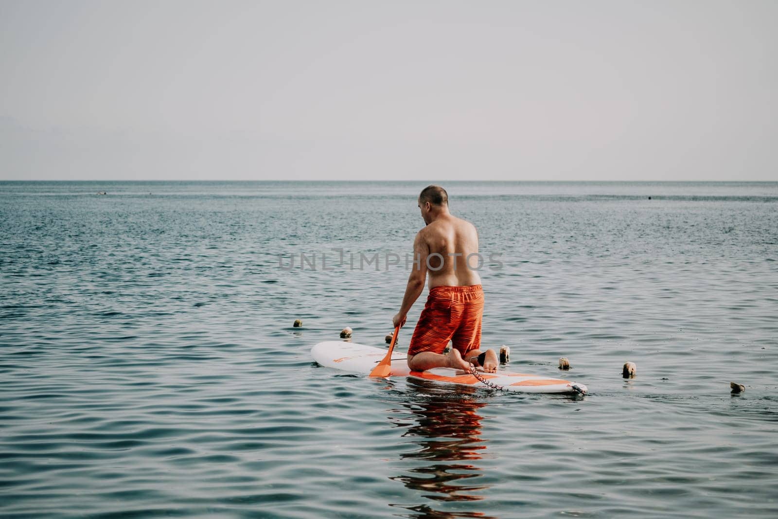 Active mature male paddler with his paddleboard and paddle on a sea at summer. Happy senior man stands with a SUP board. Stand up paddle boarding - outdor active recreation in nature