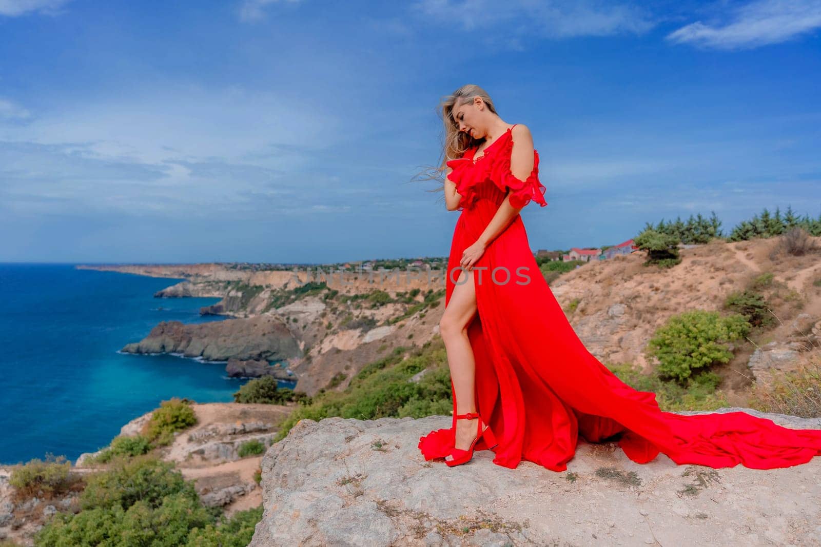 A woman in a red silk dress stands by the ocean, with mountains in the background, as her dress sways in the breeze