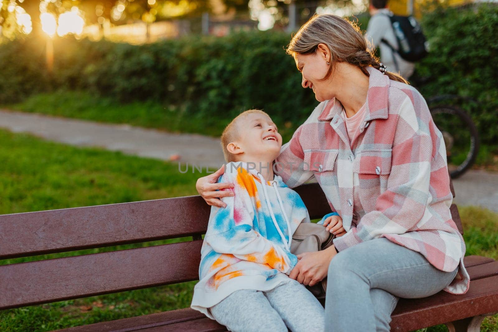 mother and son sit on a park bench in the rays of the setting sun. the concept of a family. Mother's Day. beautiful girl (mother) with a boy (son) in the park in the park are sitting on a bench at sunset by Andrii_Ko