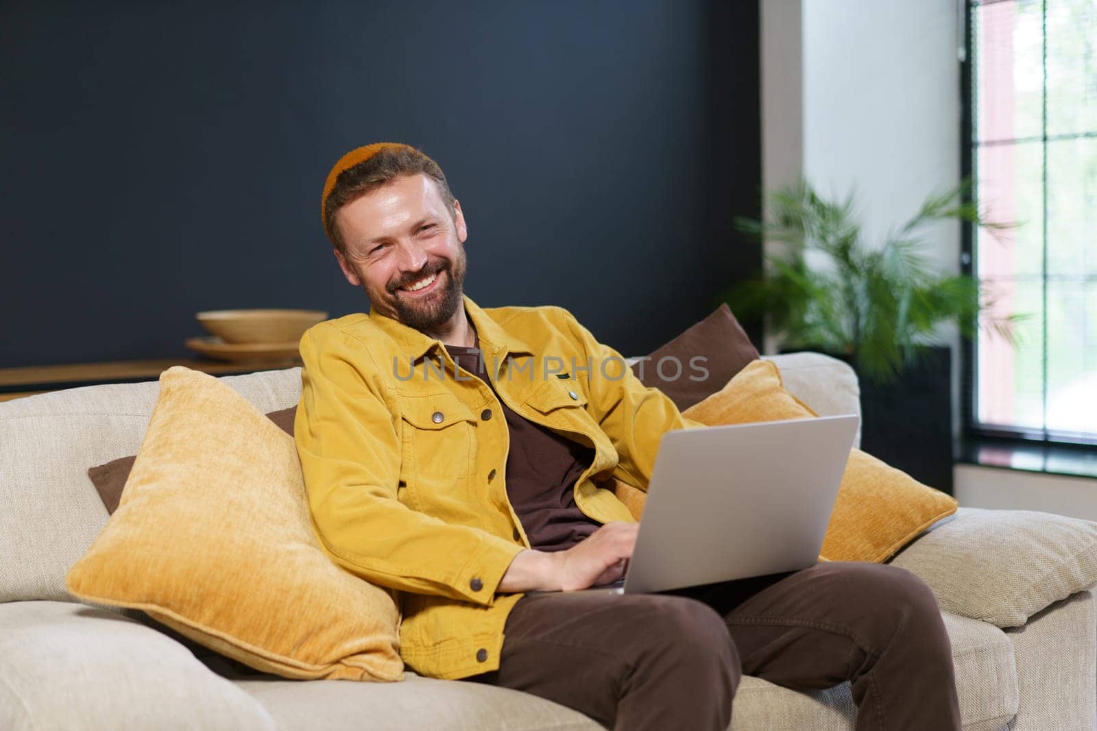 Happy and productive work-from-home scenario. Young man is seen sitting comfortably on sofa, smiling contentedly as he works on his laptop. High quality photo