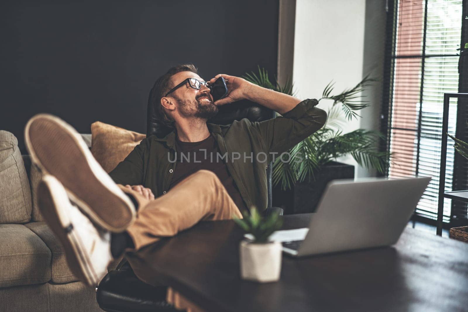 Happy and relaxed freelancer is working from home and taking phone call while sitting at desk with feet up like cowboy. He looks out window with smile on face, enjoying flexibility of remote work and comfort of his own home. by LipikStockMedia