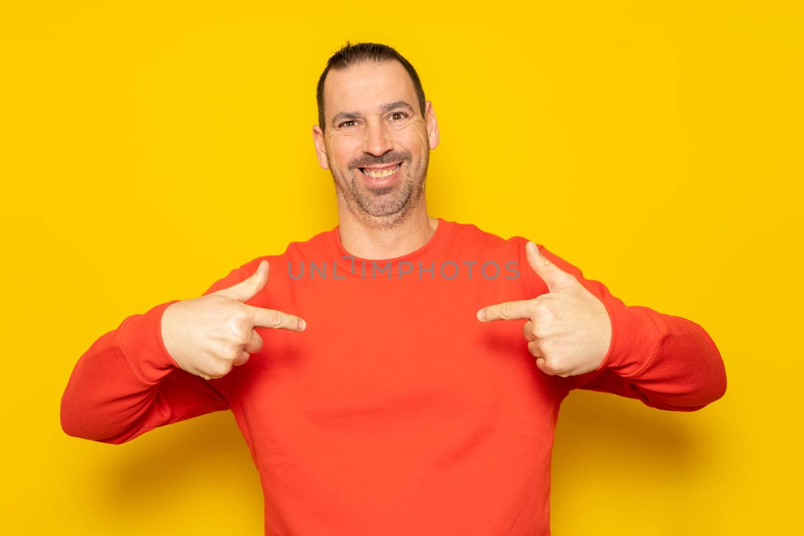 Bearded hispanic man in his 40s wearing a red jumper looking confident with a smile on his face, pointing fingers to himself proud and happy. Isolated on yellow background